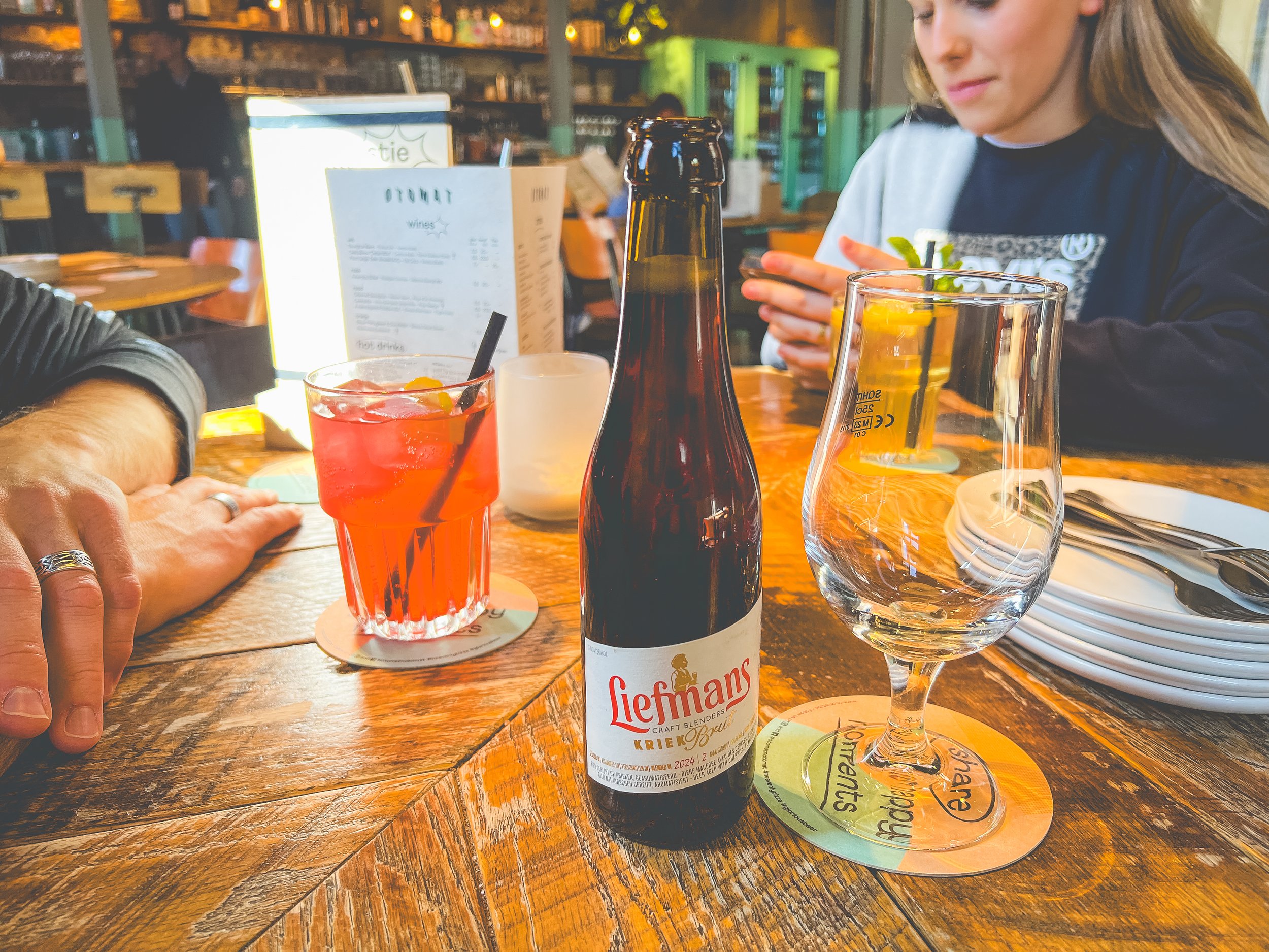 A wooden table in a restaurant with drinks including an orange beverage in a glass with a black straw, a clear empty glass, and a beer bottle labeled 'Liefmans.' There are plates, utensils, a stack of plates, and a person wearing a black shirt on the