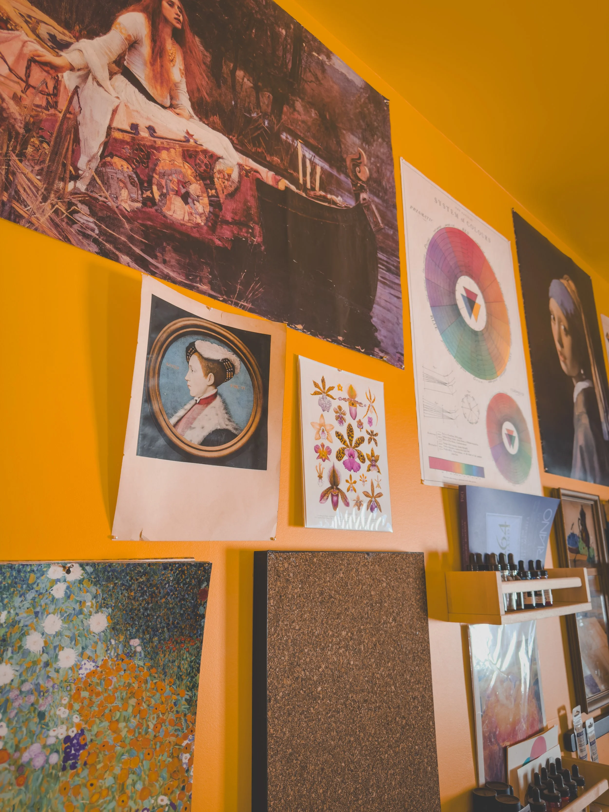 A yellow wall decorated with various posters, artwork, and charts, including a landscape painting, a historical portrait, a floral illustration, a color wheel, and a woman with a headscarf. Lady of Shallot, Girl With A Pearl Earring posters. 
