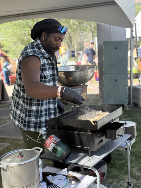 A man cooking food on a portable grill at an outdoor event, with a black and white checkered sleeveless shirt, sunglasses, and a black bandana.