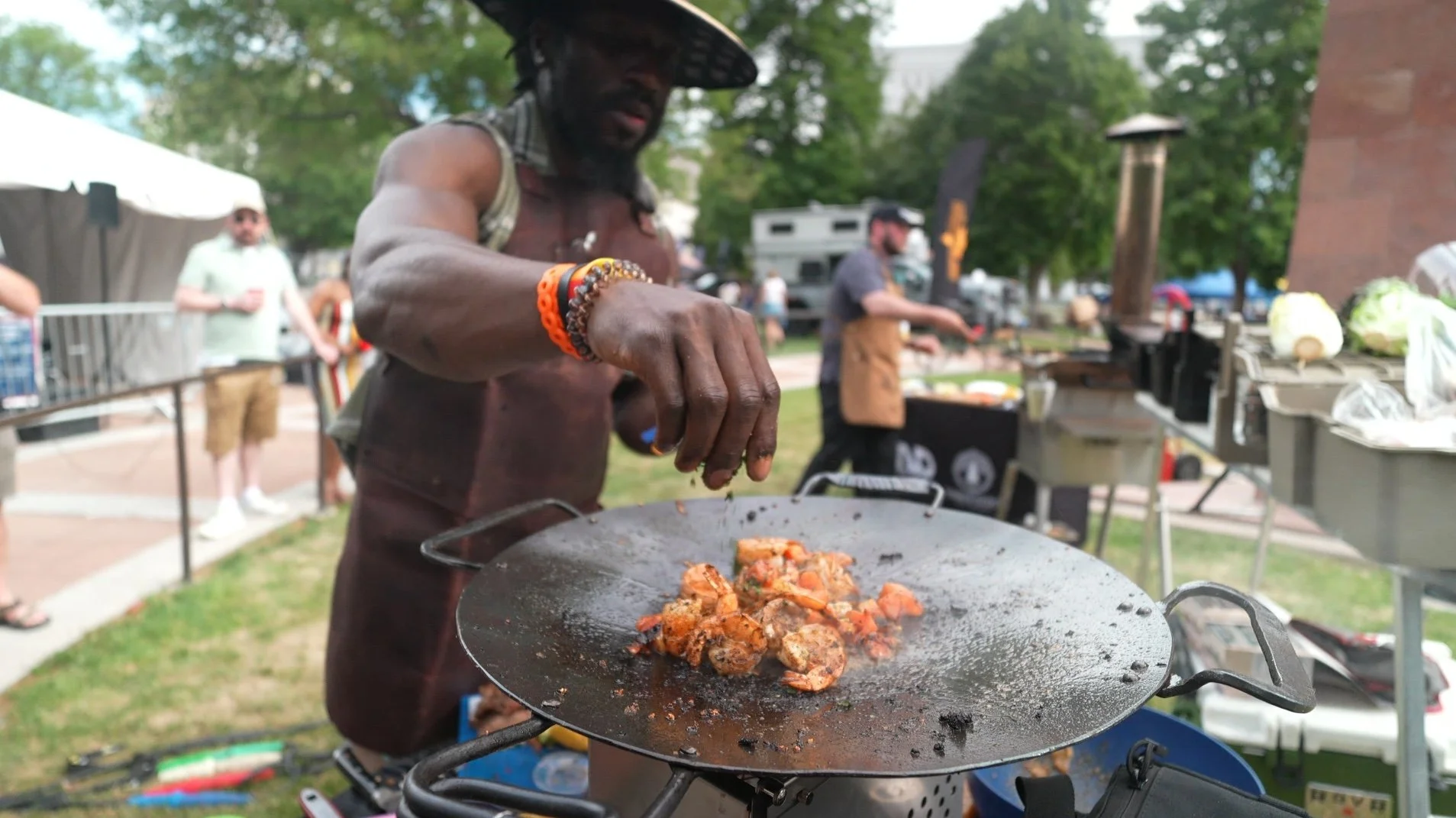 A man cooking food on a large flat grill at an outdoor event, with other people and food stalls in the background.