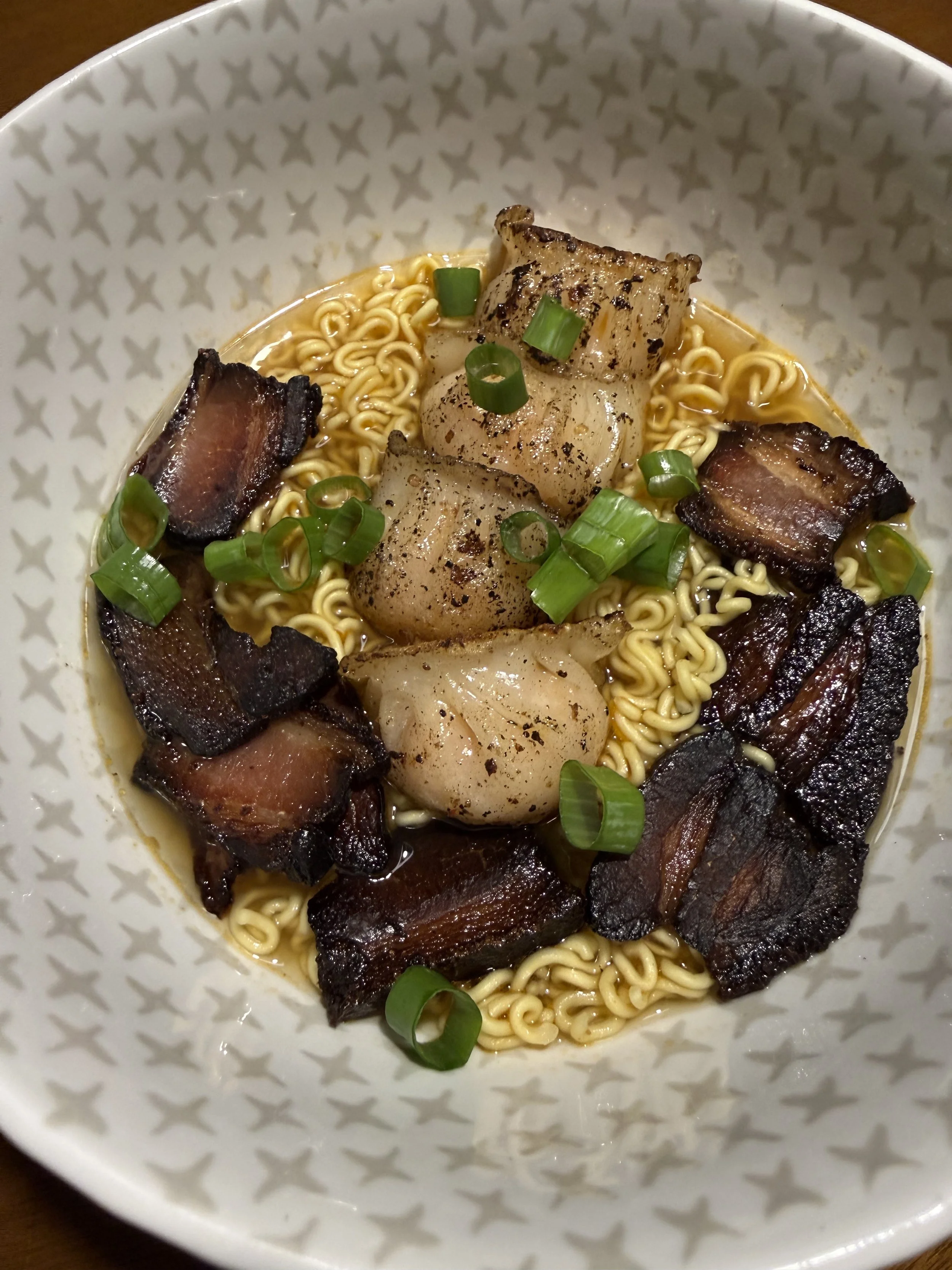 A bowl of ramen noodles topped with grilled pork belly and three seared scallops, garnished with chopped green onions.