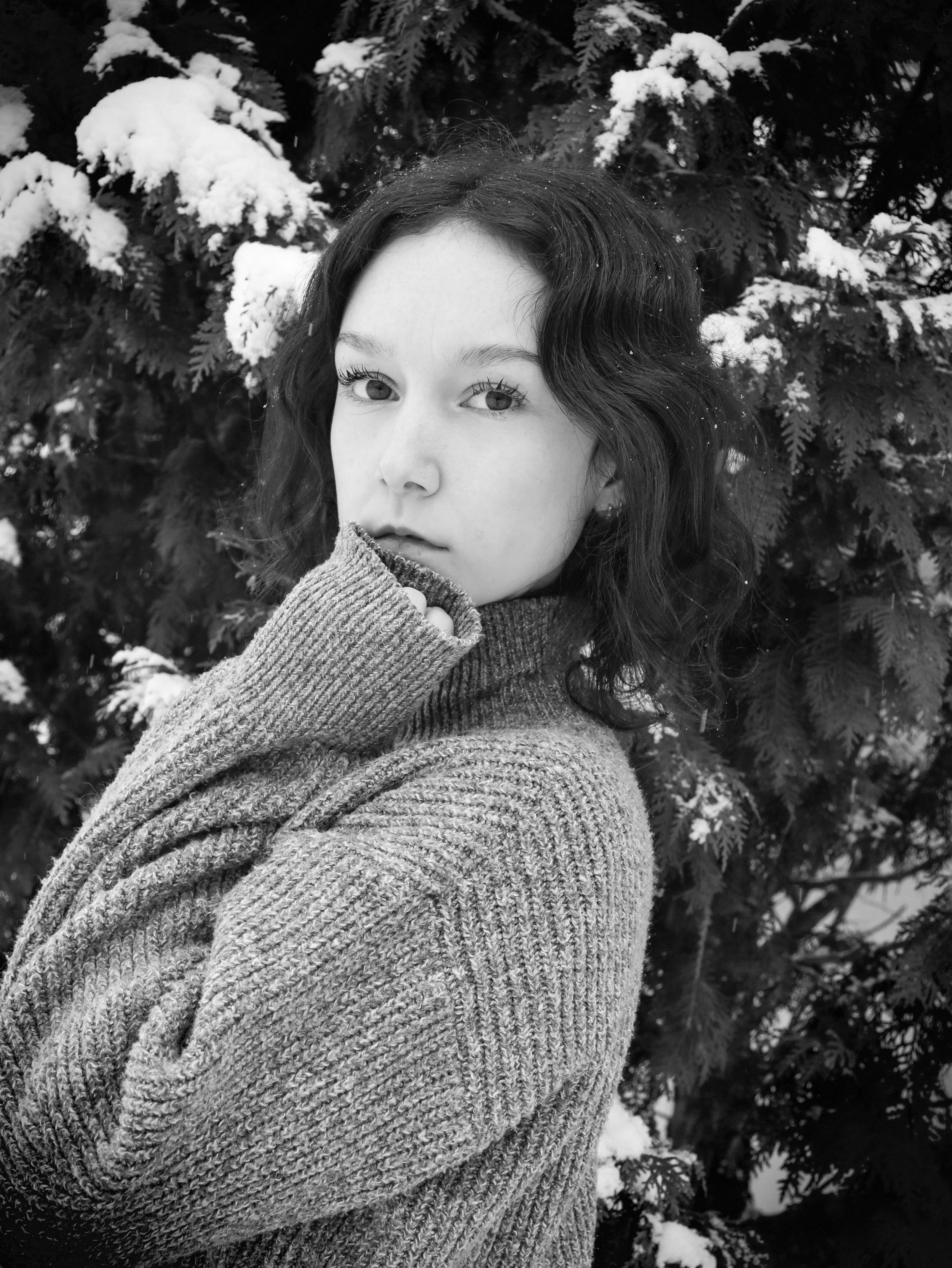 Black and white photo of a young woman with wavy hair, wearing a knitted sweater, standing in front of snow-covered evergreen trees.