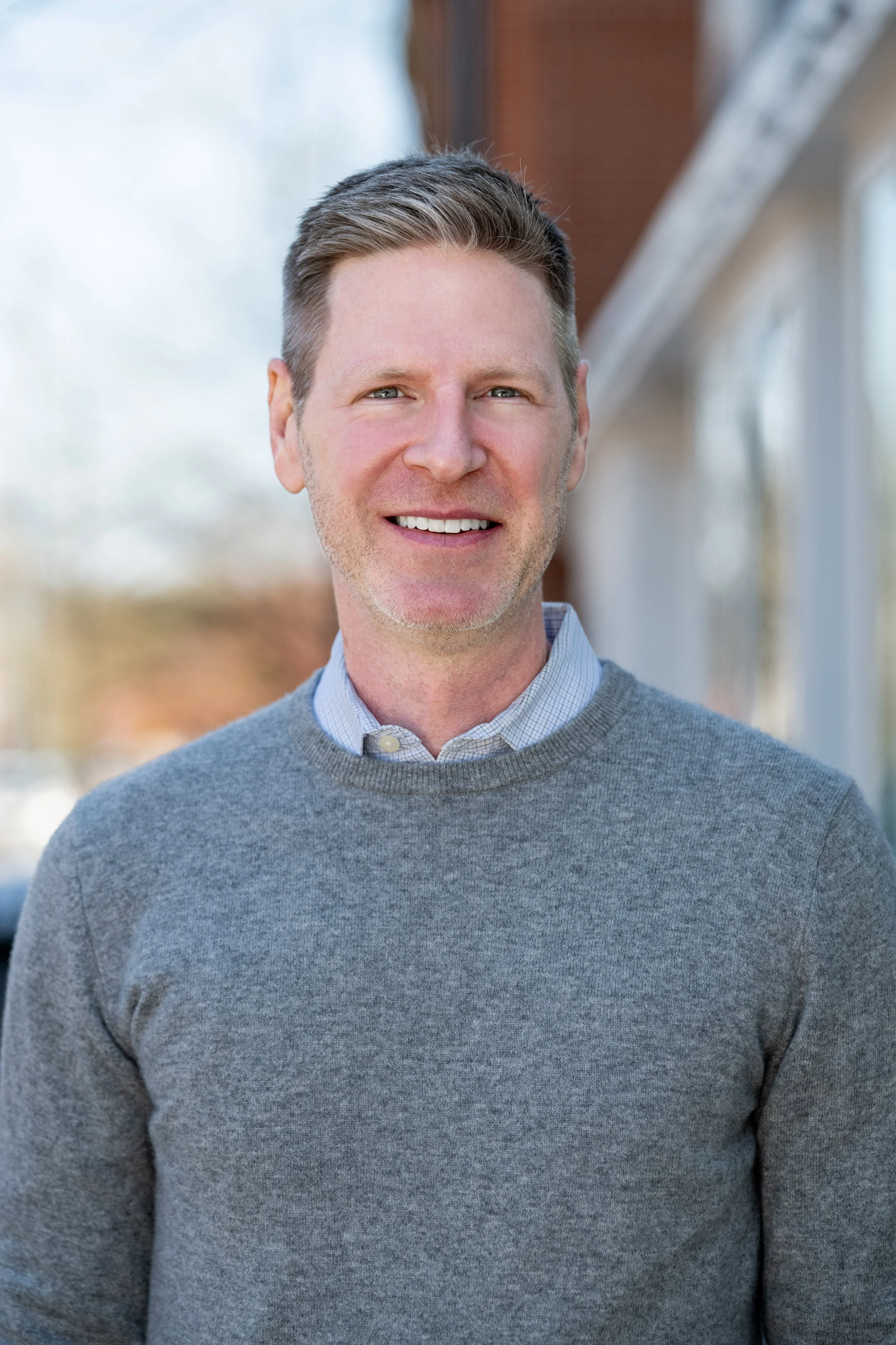 Portrait of a smiling man with short brown hair, wearing a gray sweater over a light blue collared shirt, standing outdoors with a blurred background of trees and buildings.