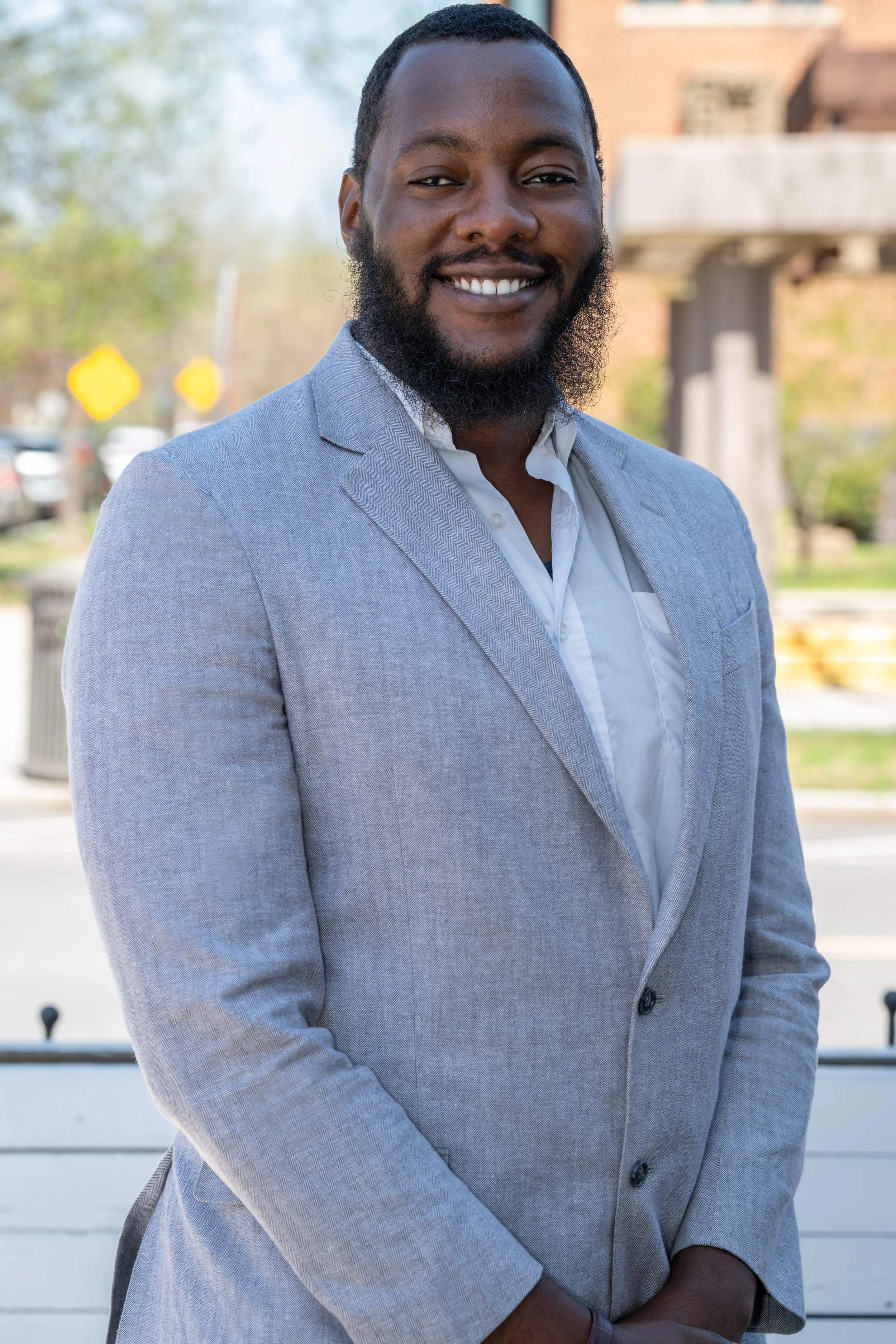 A smiling man wearing a light gray blazer and white shirt standing outdoors with trees and a building in the background.