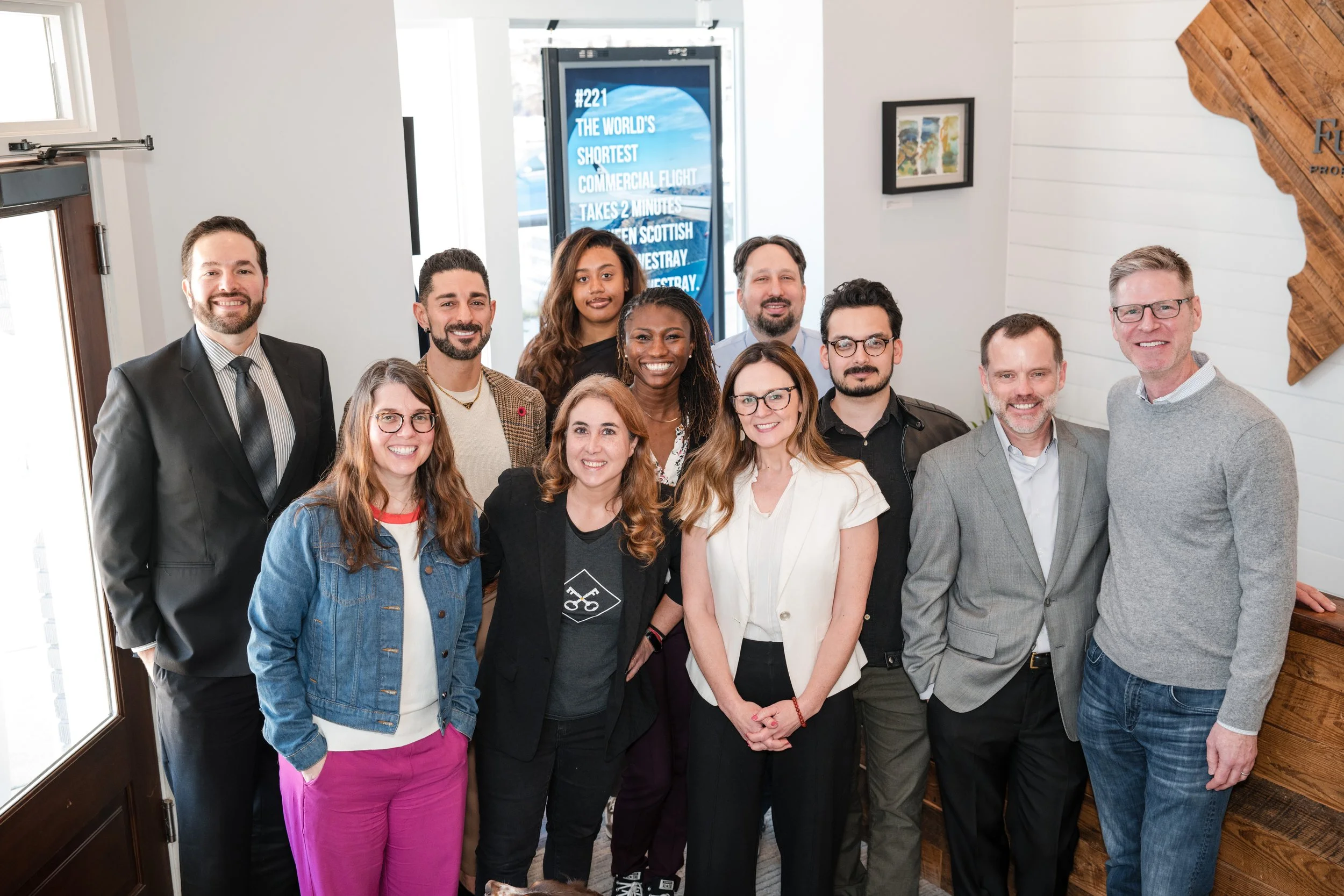 Group of twelve diverse people smiling and posing for a photo in a bright indoor setting, with a sign and artwork in the background.