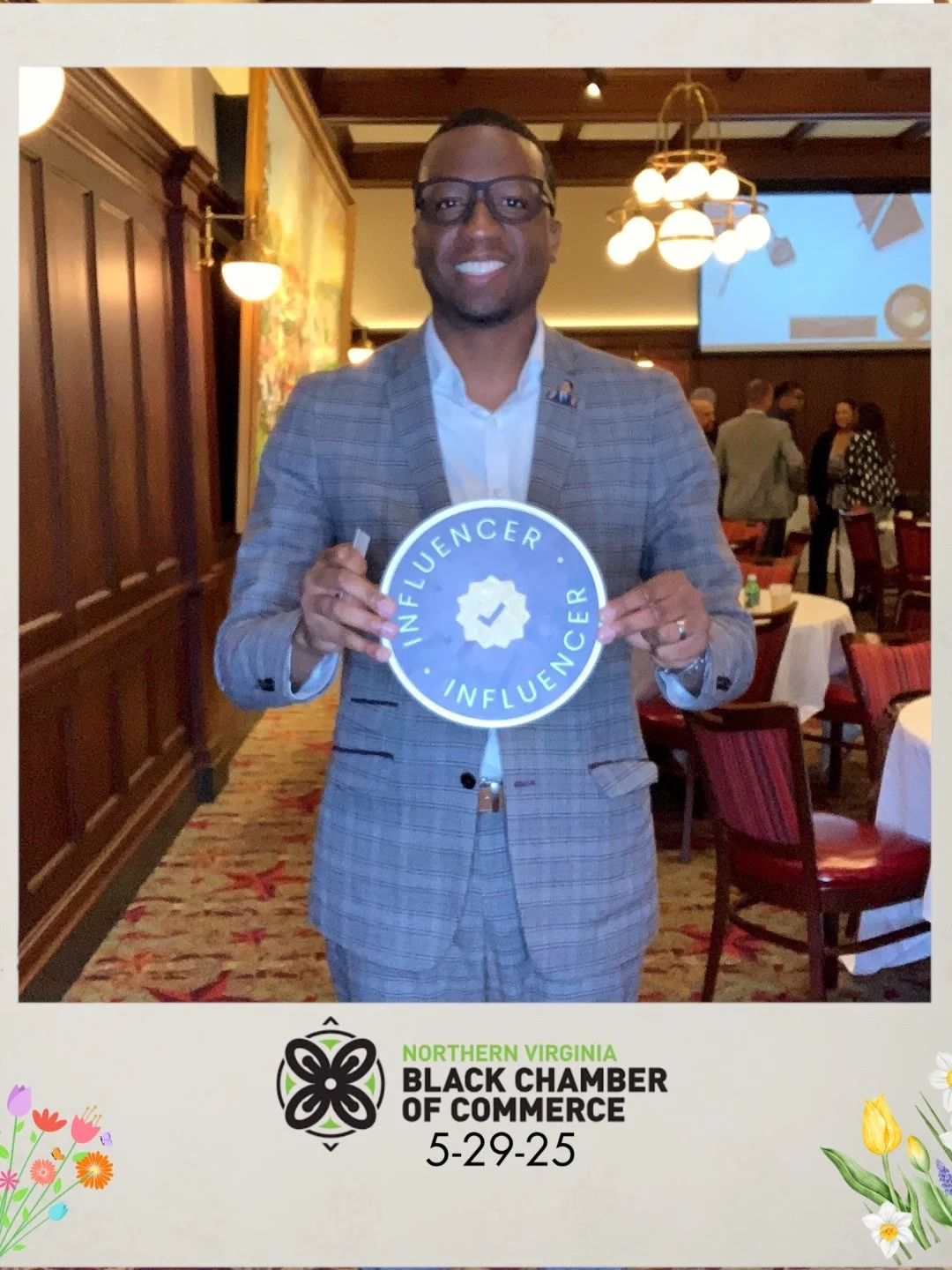 A man in a gray checkered suit holding an 'influencer' sign at a Northern Virginia Black Chamber of Commerce event on May 29, 2025, in a restaurant or banquet hall with wooden walls, round tables with white tablecloths, and other people in the background.