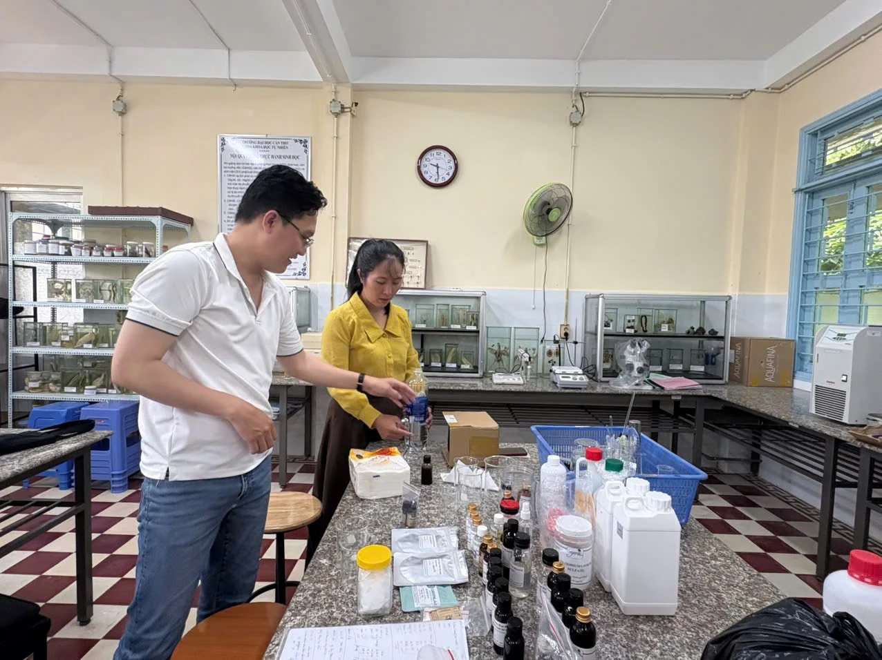 Two people working in a laboratory with various bottles and equipment on a granite table.