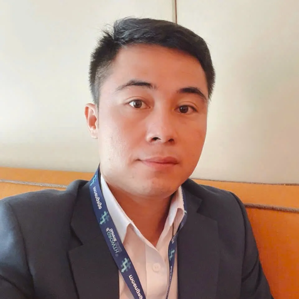 A young man in professional attire sitting at a table, wearing a suit jacket, white shirt, and a lanyard, with a neutral expression and short black hair.