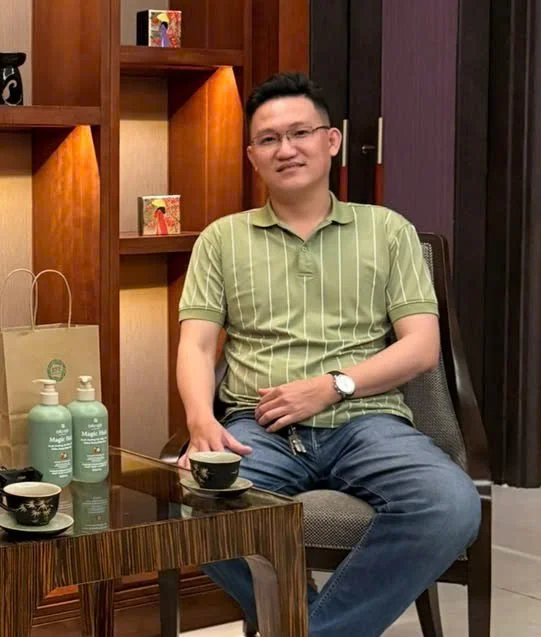 A man sitting on a chair at a table with tea cups, bottles of lotion, a paper bag, and decorative items on wooden shelves behind him.