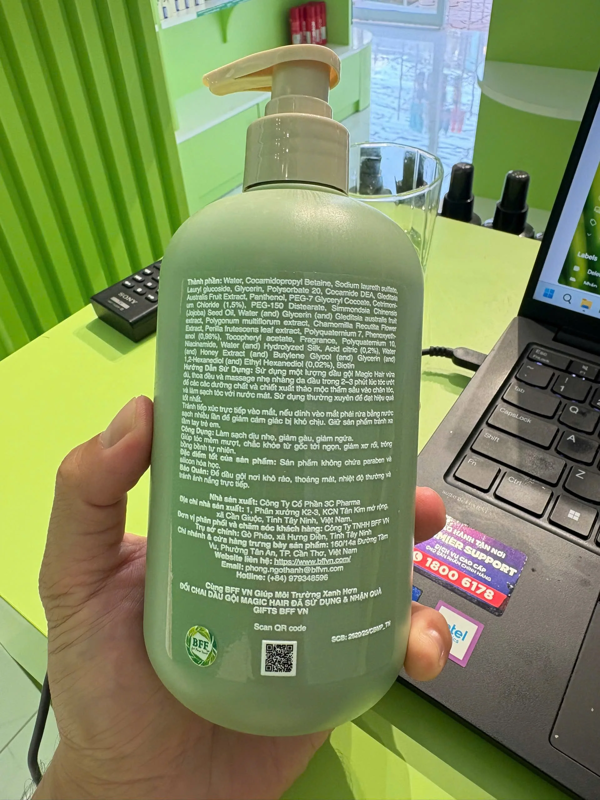 A hand holding a green bottle with a pump dispenser, showing text in Vietnamese on the back. The background includes a computer keyboard, a glass of water, and part of a green-themed workspace.