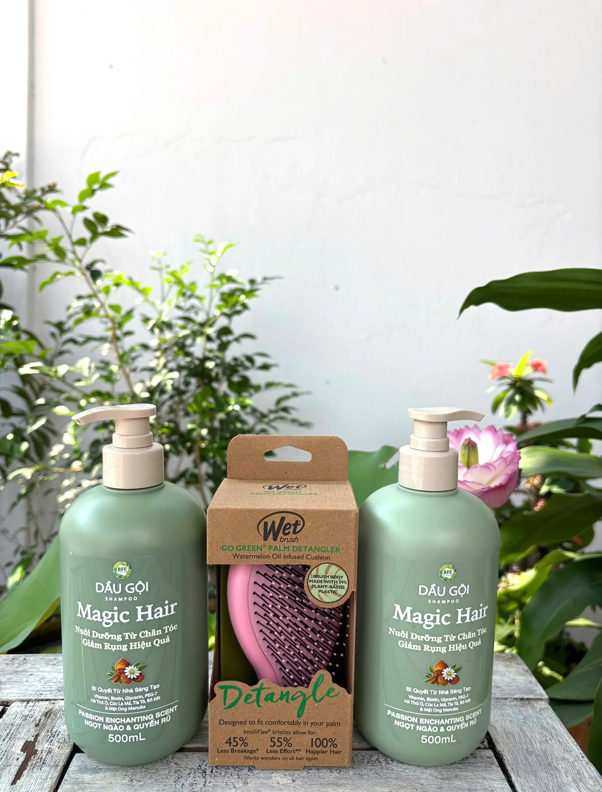 Two bottles of shampoo labeled 'Magic Hair' and a pink detangling brush in a cardboard box, set on a wooden surface with green plants in the background.