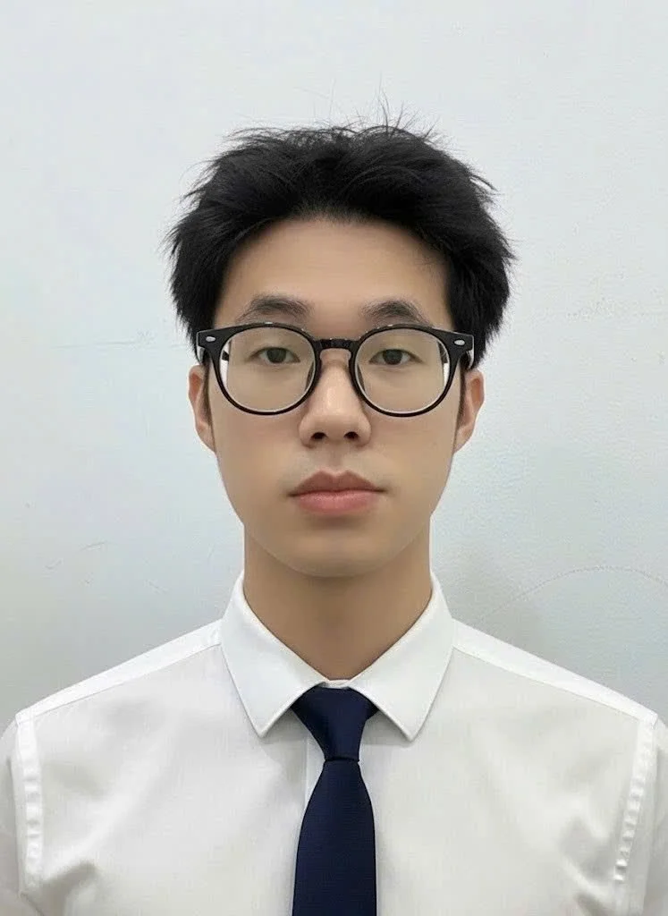 A young man with black hair, wearing black-rimmed glasses, a white dress shirt, and a navy blue tie, standing against a plain light-colored background.