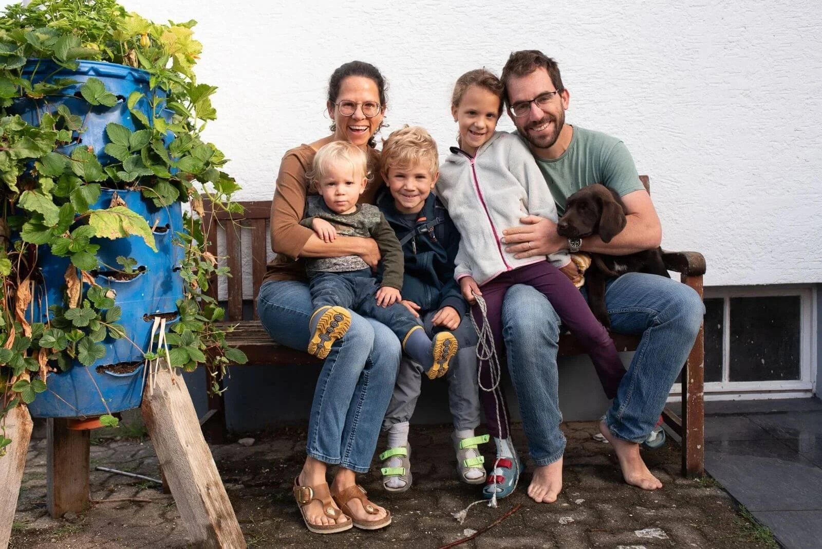 Familie mit drei Kindern und Hund sitzt auf einer Holzbank vor dem Werkstattgebäude