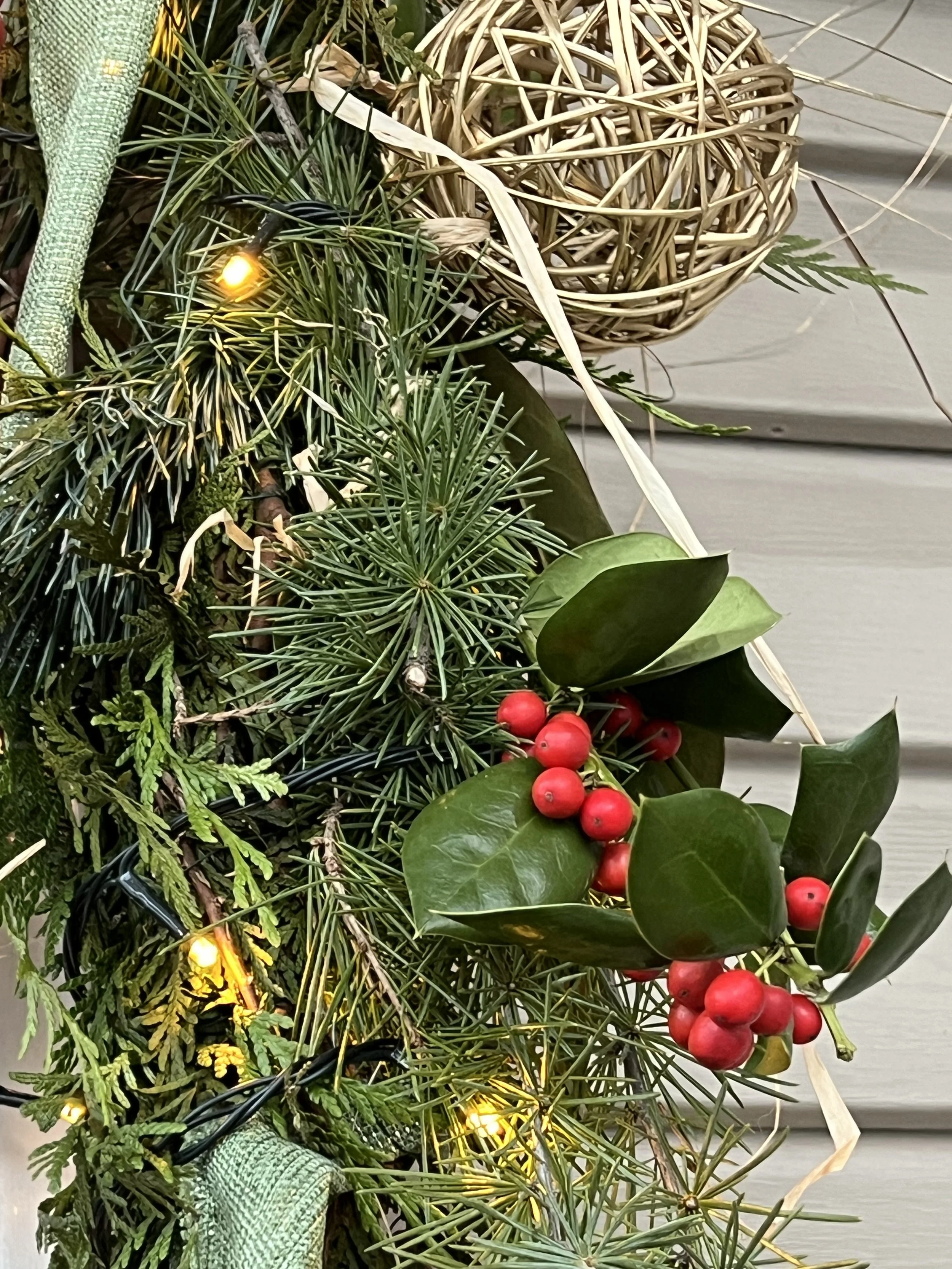 Close-up of a Christmas decoration with green pine, holly leaves, red berries, a woven rattan ball, and yellow string lights in Annapolis, MD.
