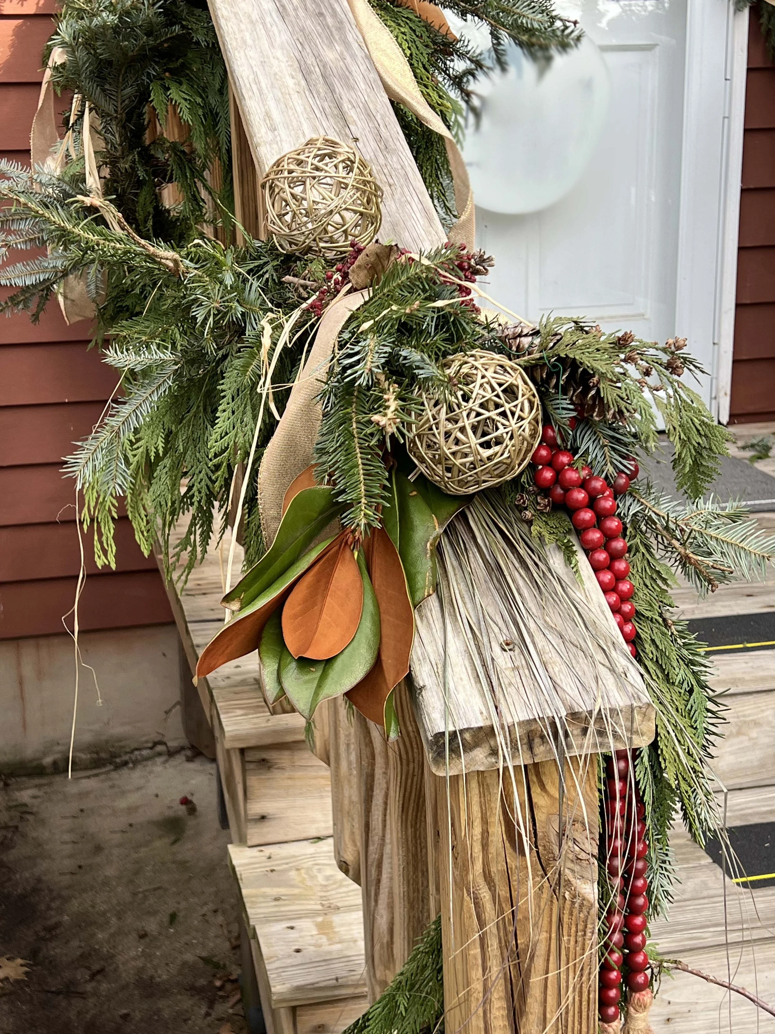A close-up of a rustic Christmas or holiday decoration featuring evergreen branches, dried leaves, red berries, wicker balls, and beige ribbon on a wooden railing outdoors in Annapolis, Maryland.