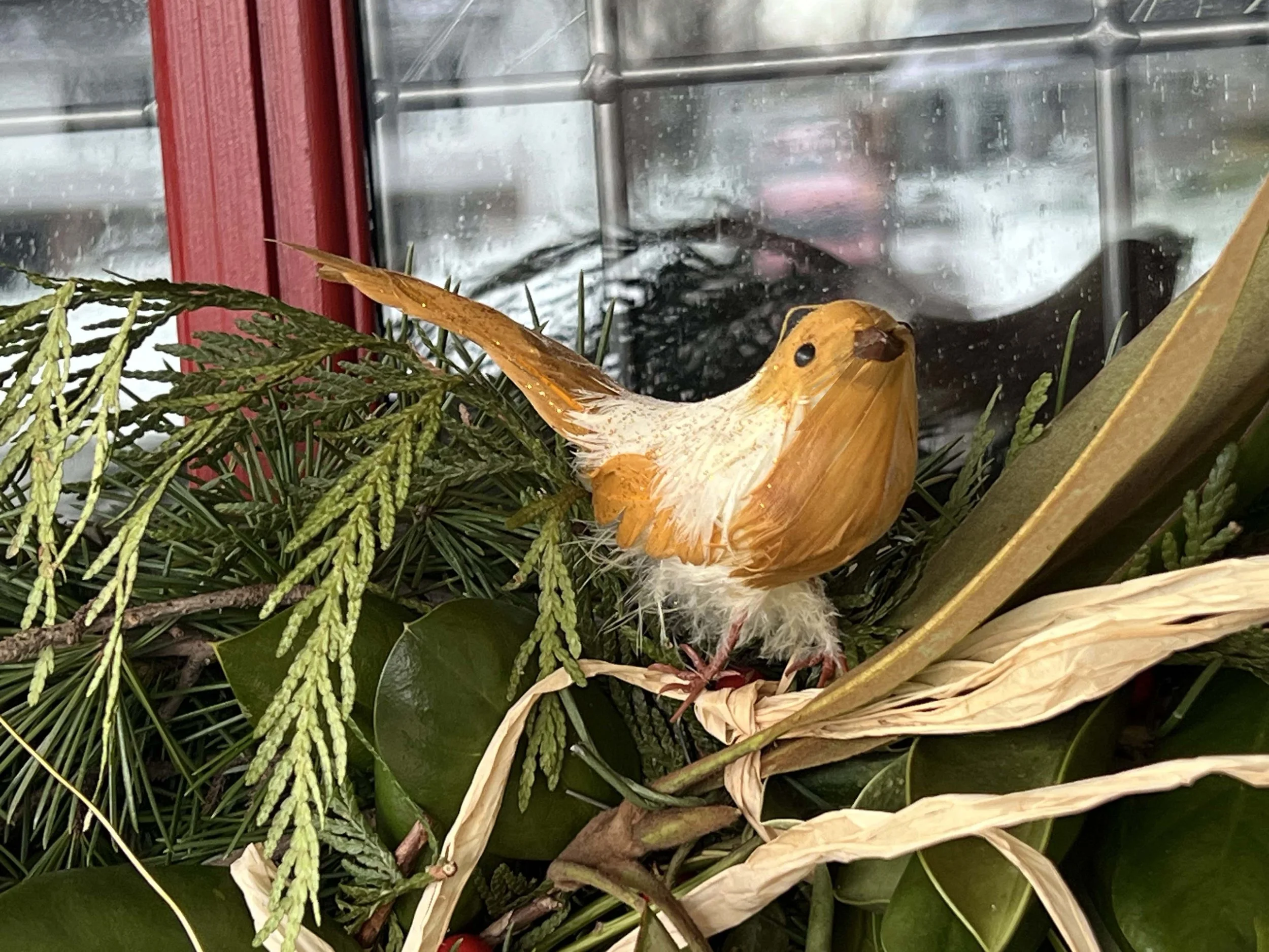 Artificial bird decoration with orange and white feathers perched among green leaves and pine branches, in front of a window with red frame. Front Door Decor Annapolis, MD