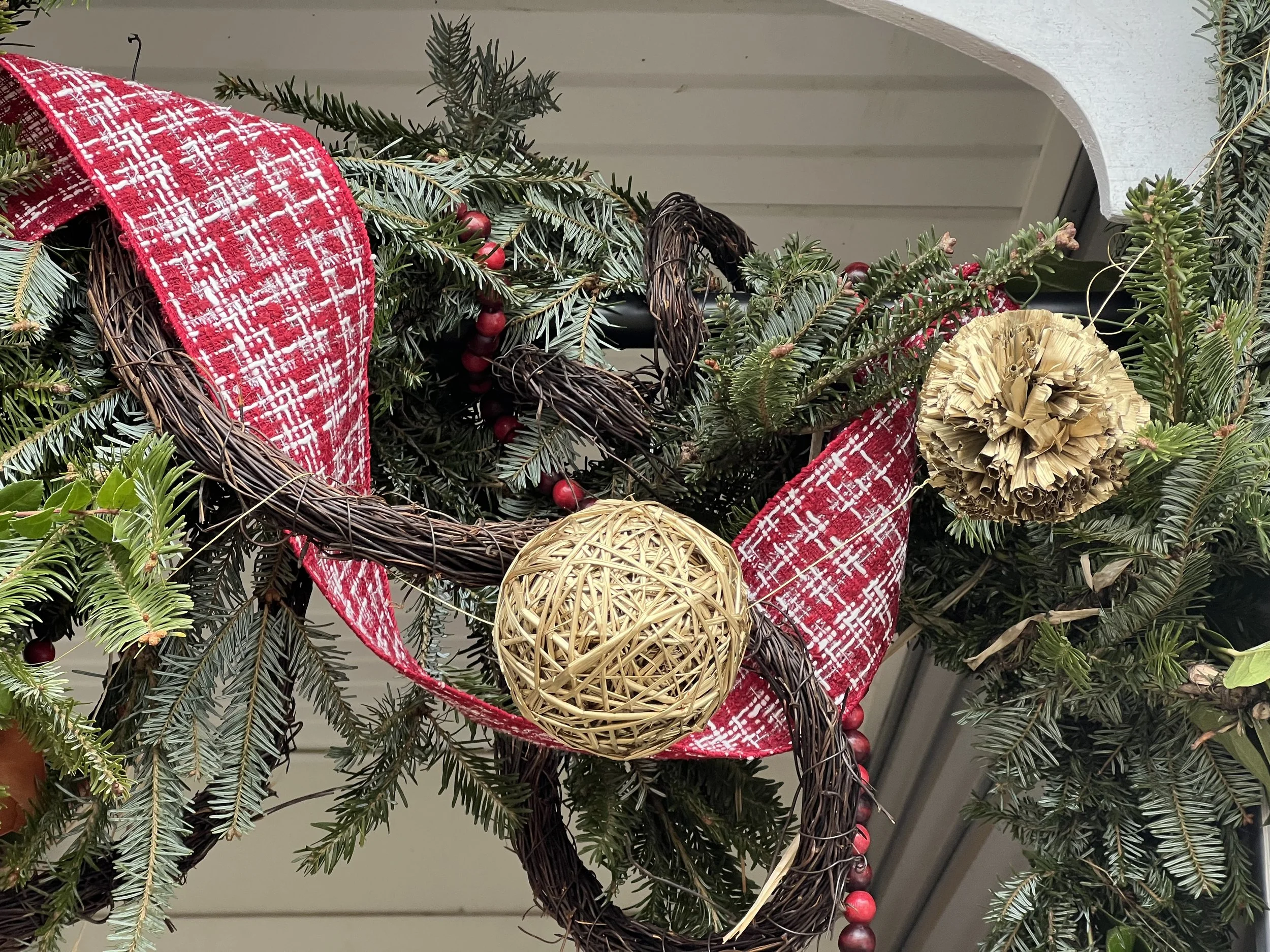 Close-up of a Christmas wreath decorated with red plaid ribbon, red berries, a woven beige ball, and a woven gold ball, surrounded by green pine branches in Severna Park, Maryland.