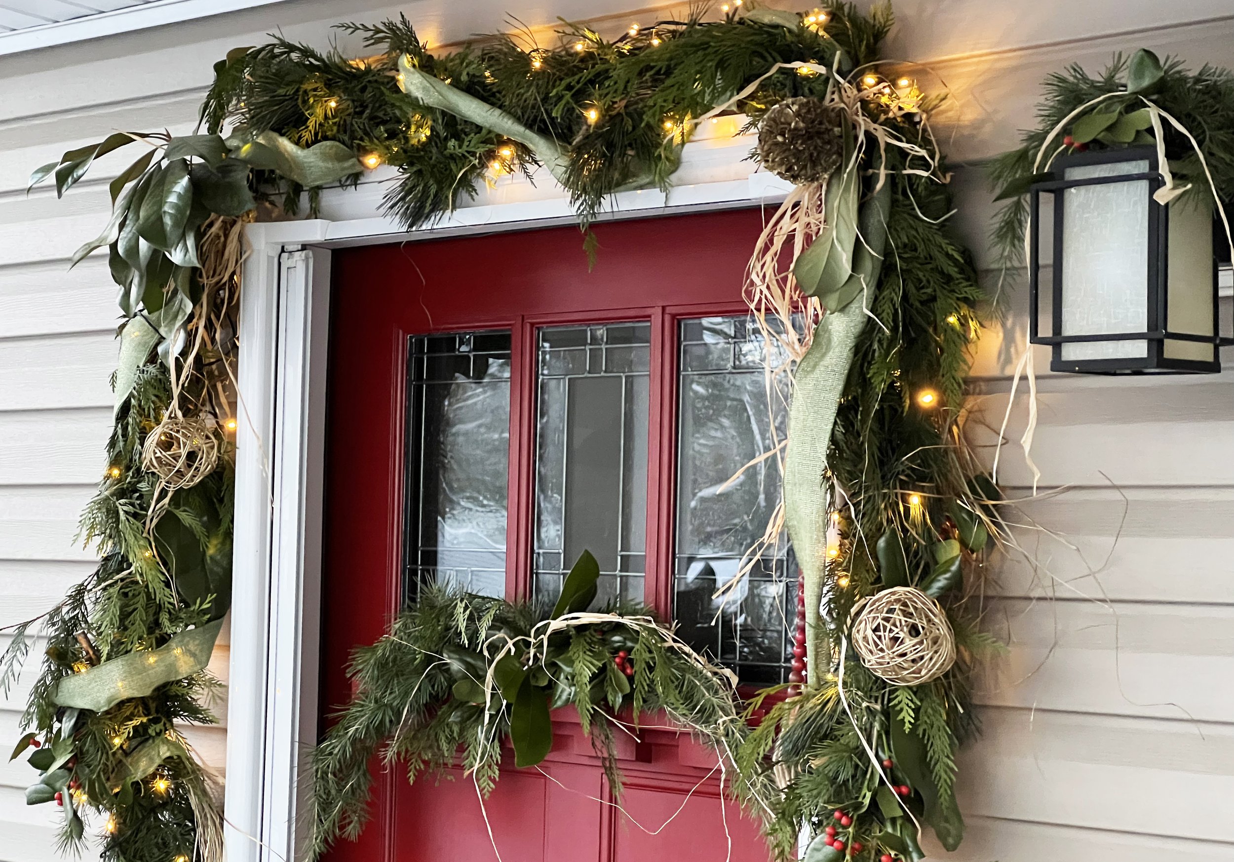 Front door decorated with a lush green Christmas Decorations with pine, holly, ribbons, and small ornaments, dressed with warm white string lights, on a house with beige siding in Annapolis, Maryland.