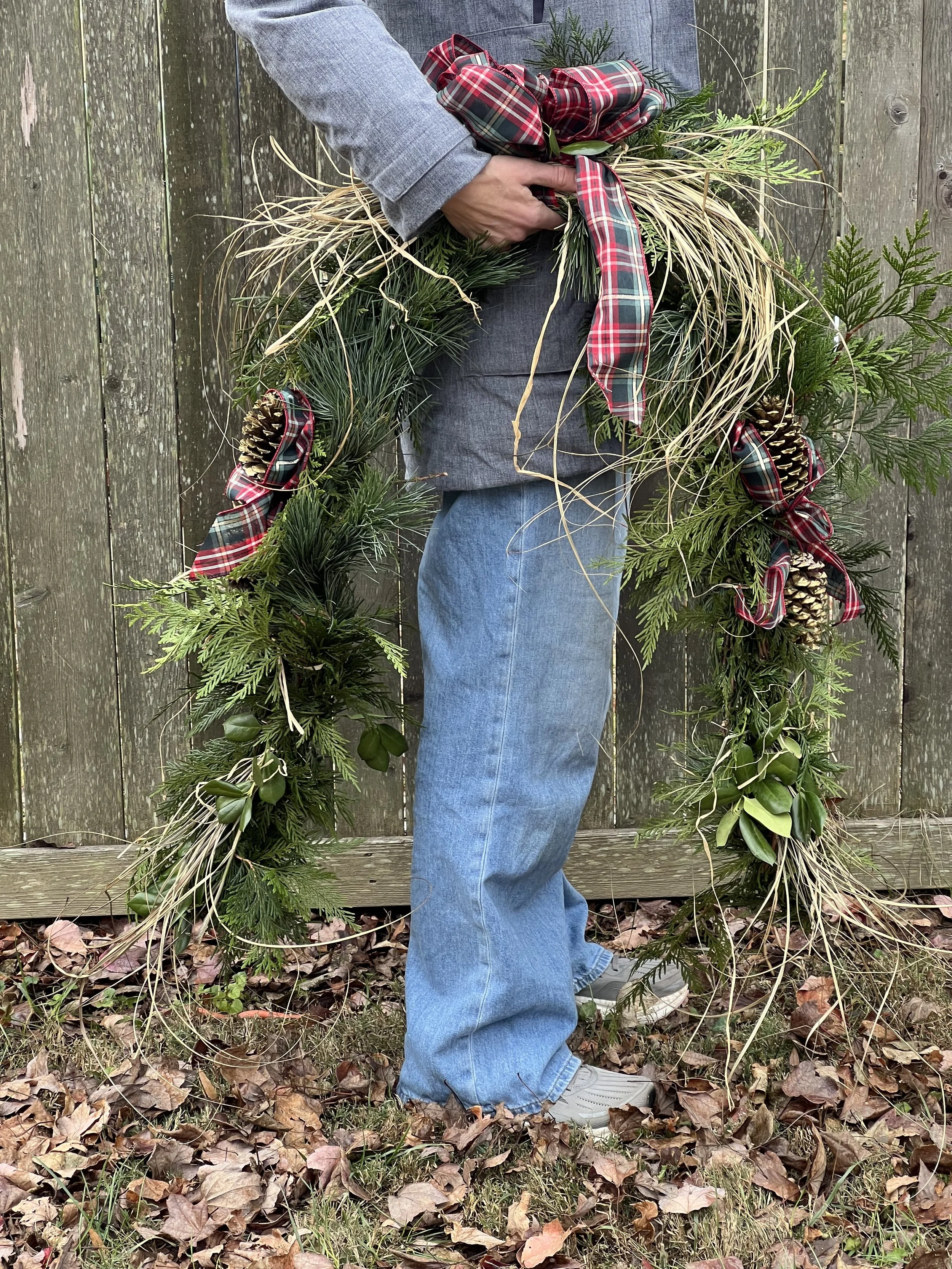 Person holding a Christmas wreath made of greenery, pinecones, and plaid ribbons outdoors in front of a wooden fence, with fallen autumn leaves on the ground in Annapolis, Maryland. 