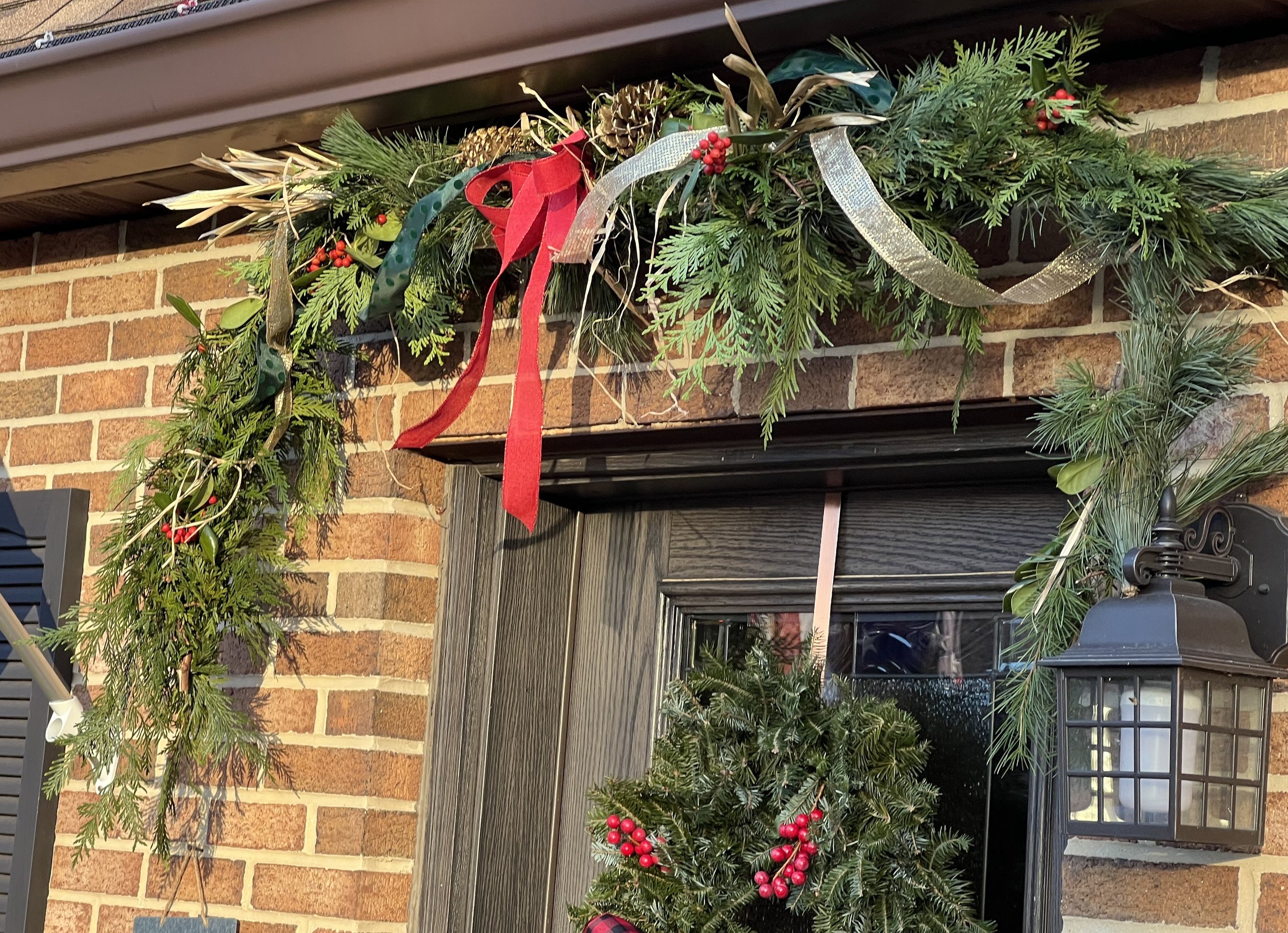 Christmas decorations with greenery, red berries, and ribbons hanging above a door with a wreath decorated with red berries, next to a black lantern on brick wall in Annapolis, Maryland. 
