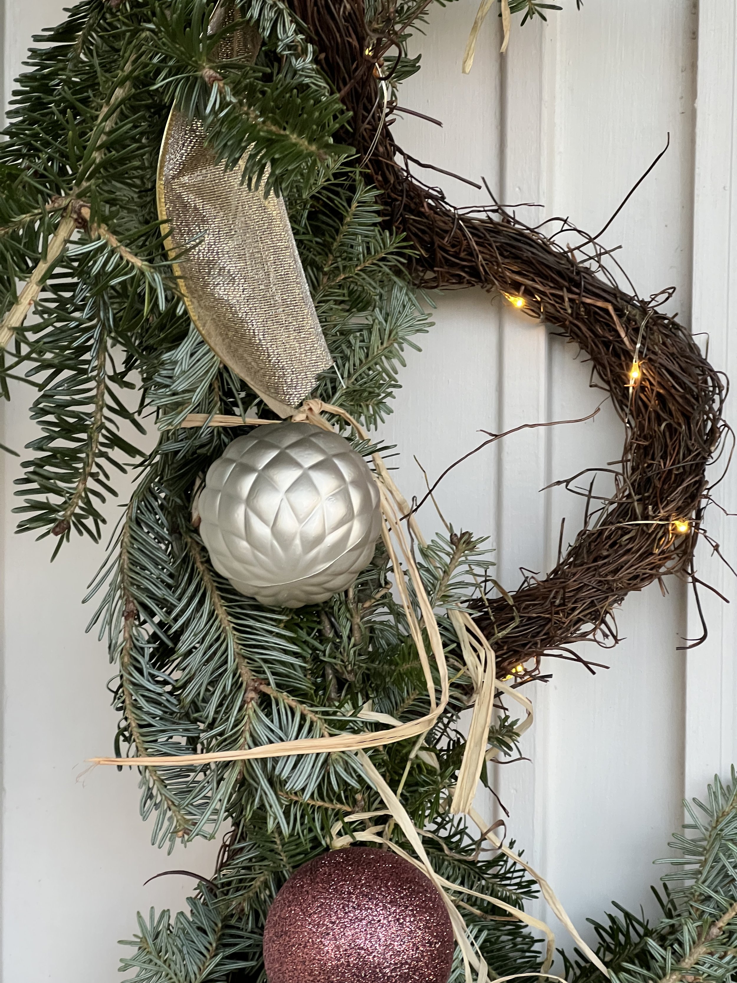 A Christmas wreath decorated with pine branches, two ornamental balls (one silver and one red), a gold ribbon, and string lights in Annapolis, Maryland.