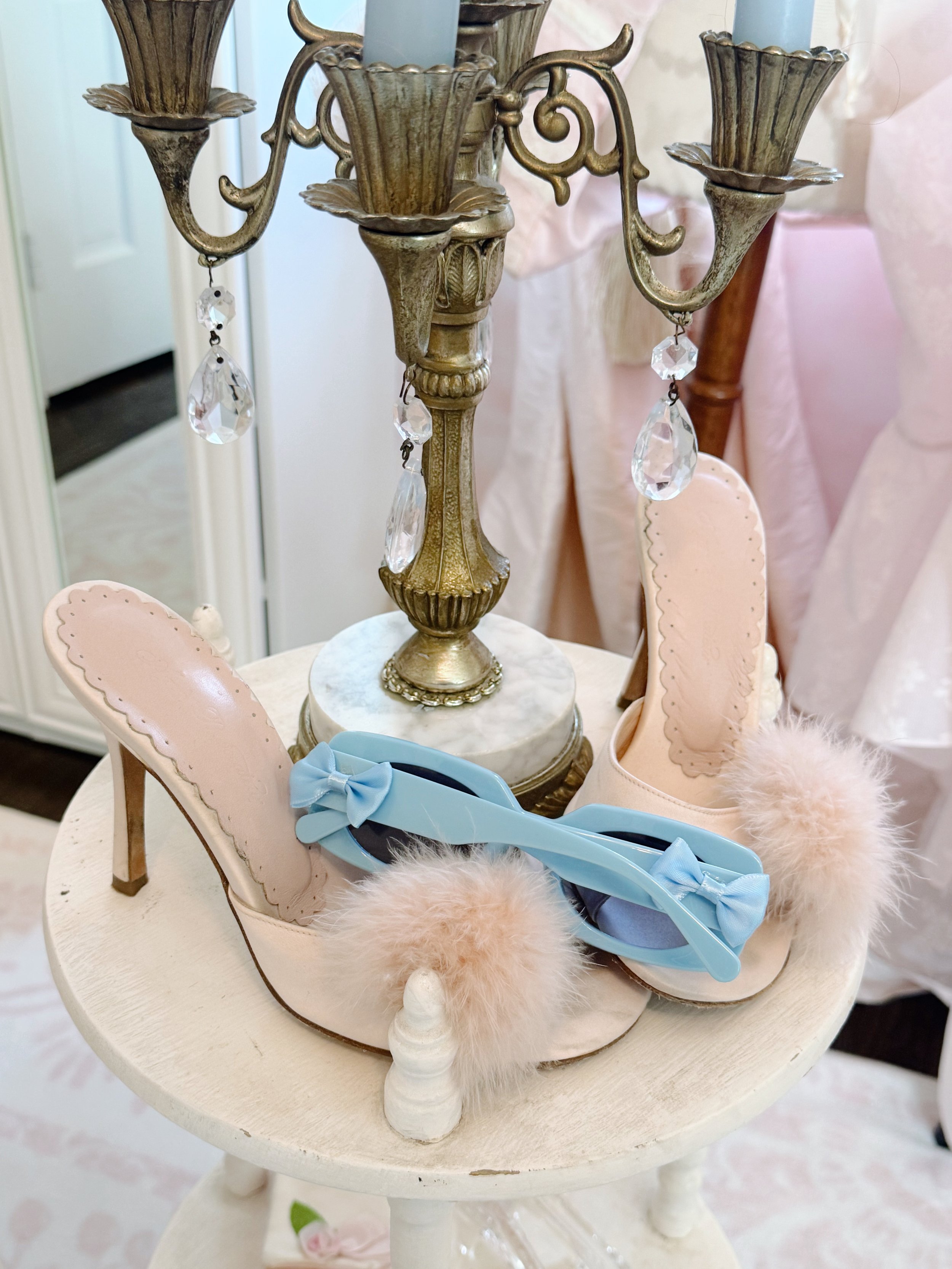 A vintage-style table holding a brass and crystal chandelier, pink high-heeled shoes with fluffy pom-poms, and a pair of light blue glasses with bows.