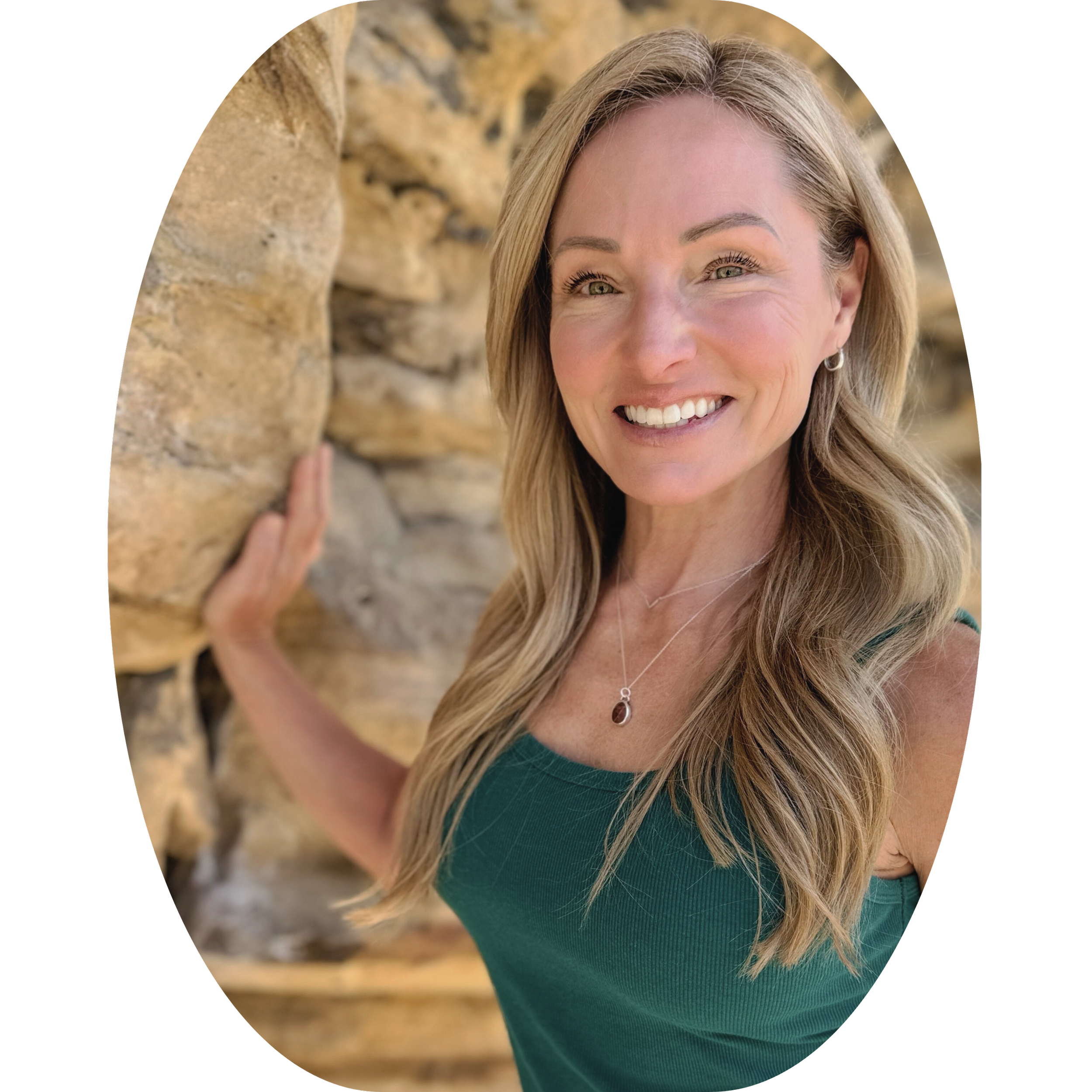 A woman with long blond hair smiling and leaning against a stone wall outdoors.