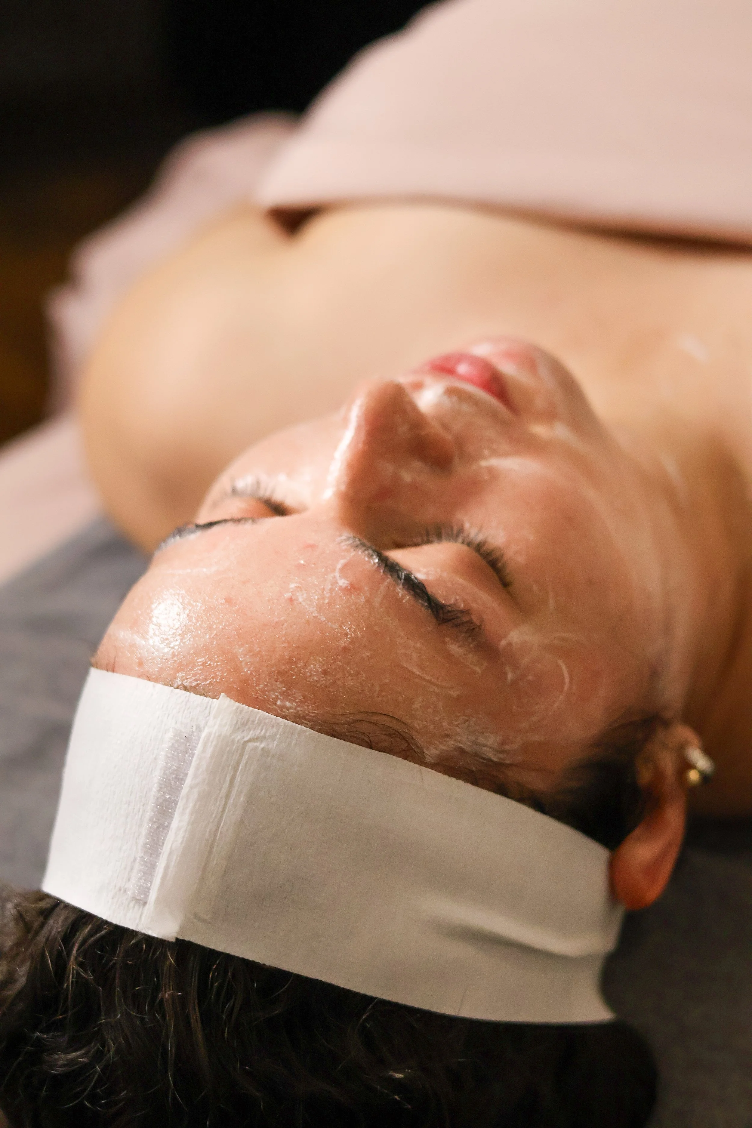 A woman lying on a treatment bed with a headband, receiving a facial treatment with a visible thick cream or mask applied to her face, in a spa or skincare setting.