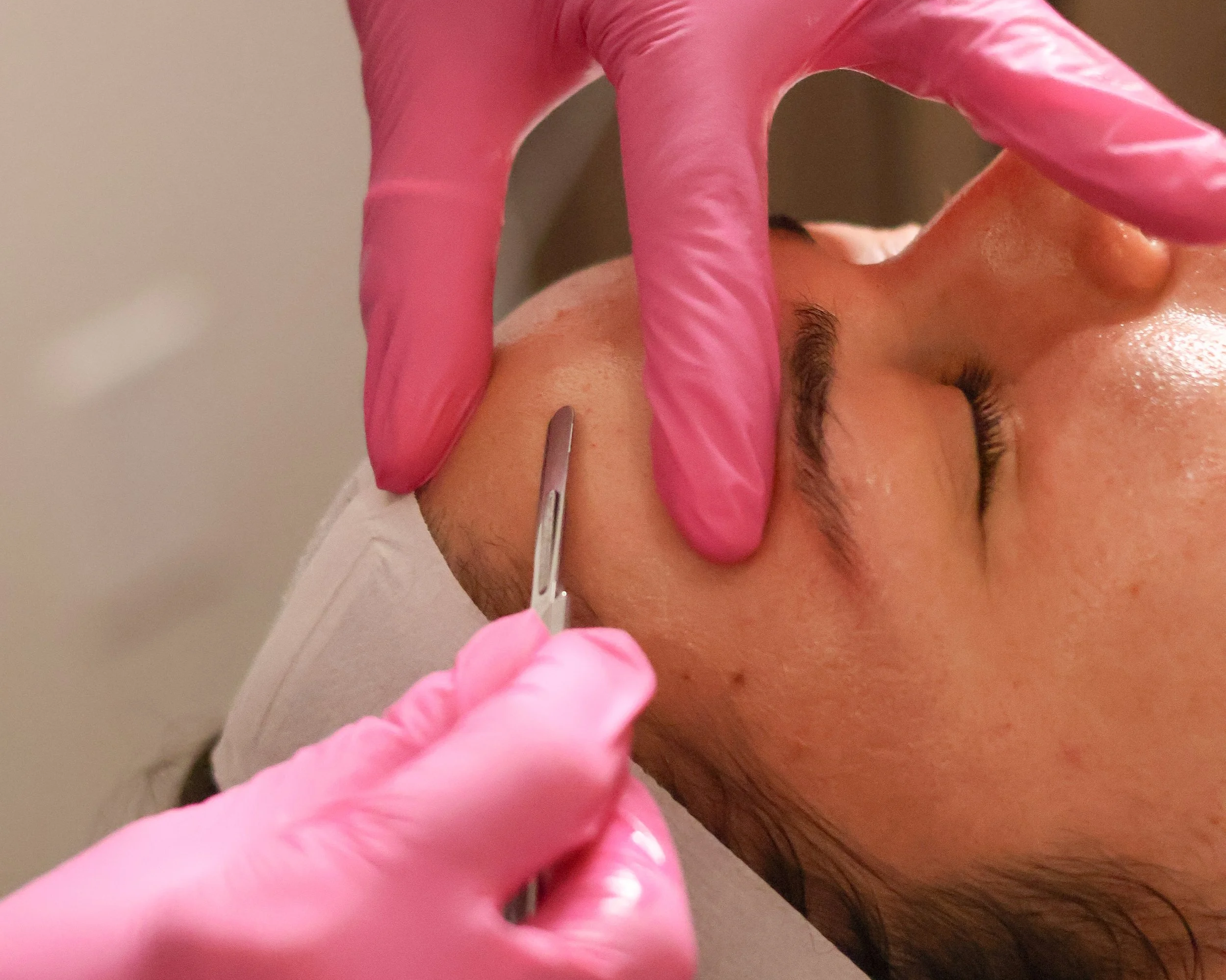 A person wearing pink gloves is performing a cosmetic procedure on a man's eyebrow using a small precision tool.