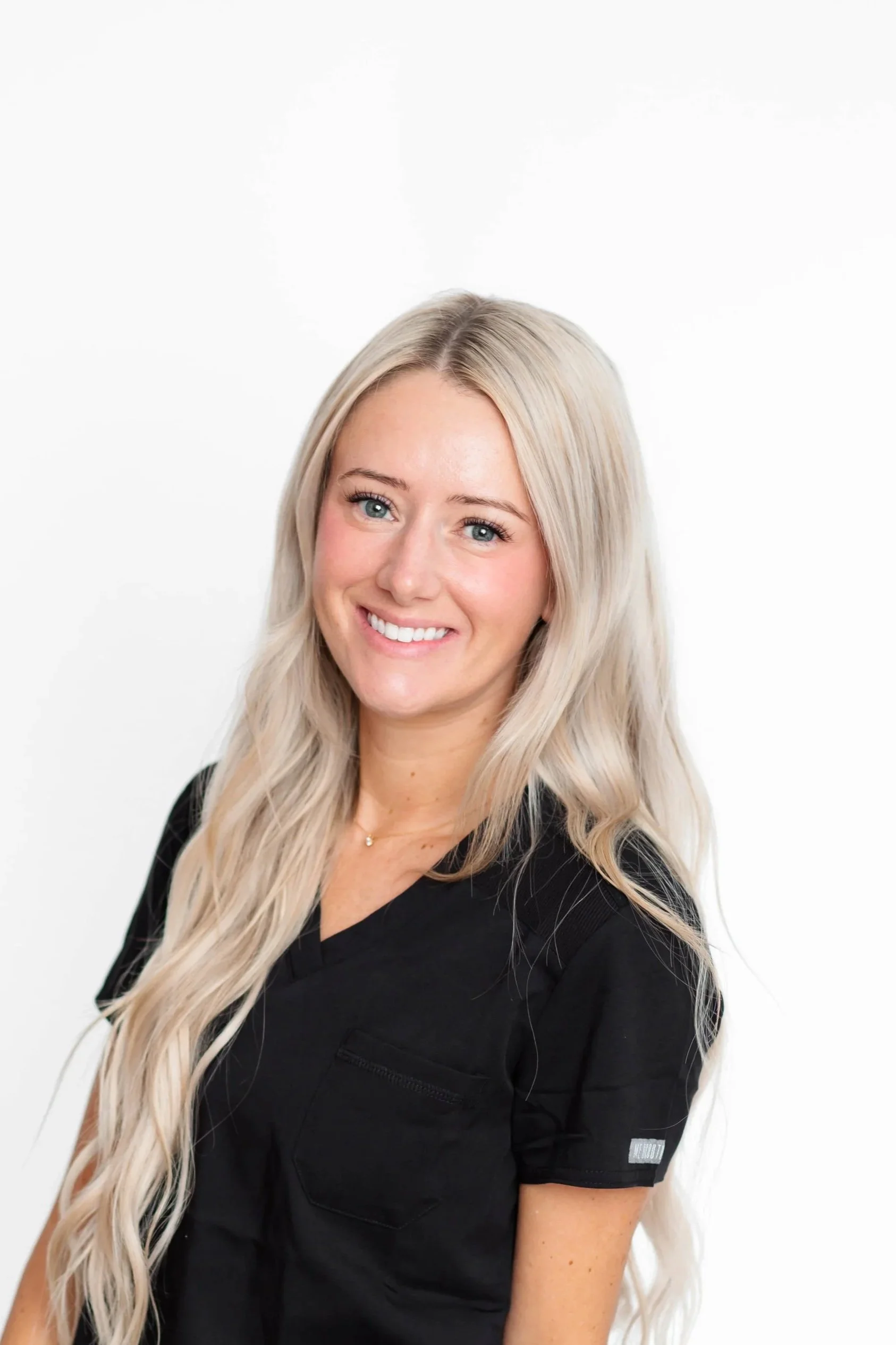 A young woman with long, wavy blonde hair smiling at the camera, wearing a black top, against a white background.