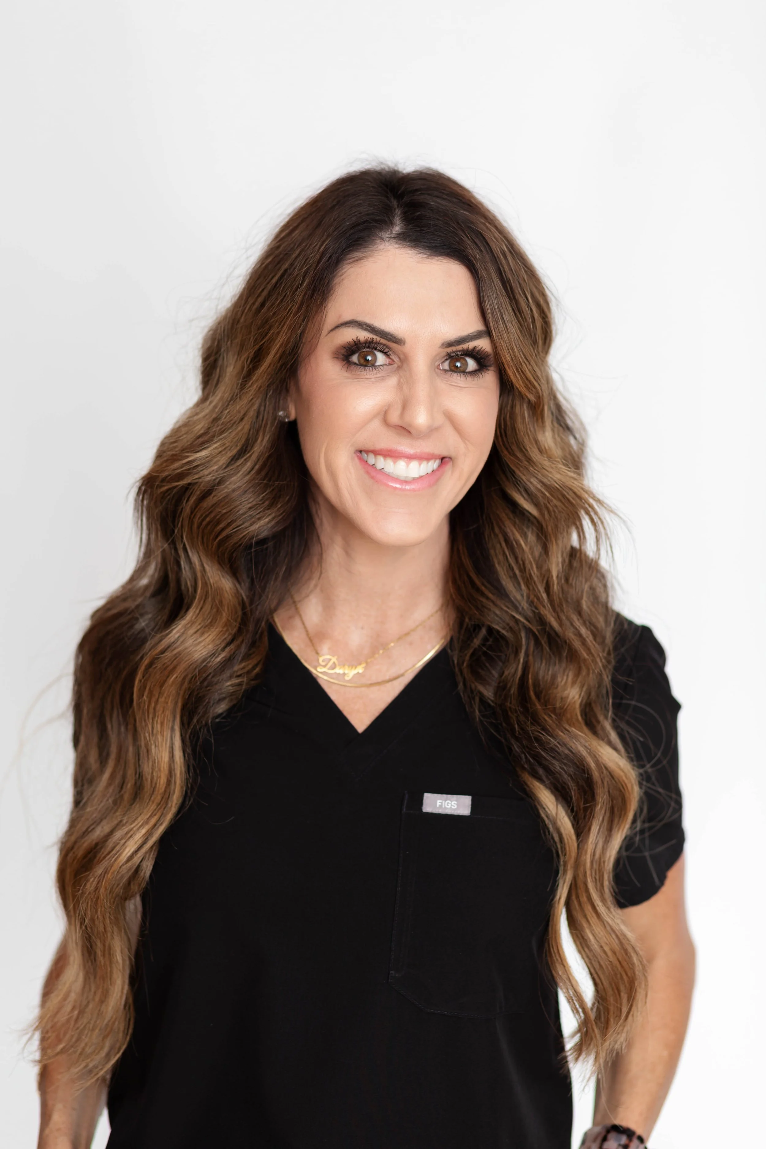 A woman with long wavy brown hair, smiling, wearing a black medical uniform with a 'FIGS' logo on the chest, and a gold necklace that says 'Dany'.