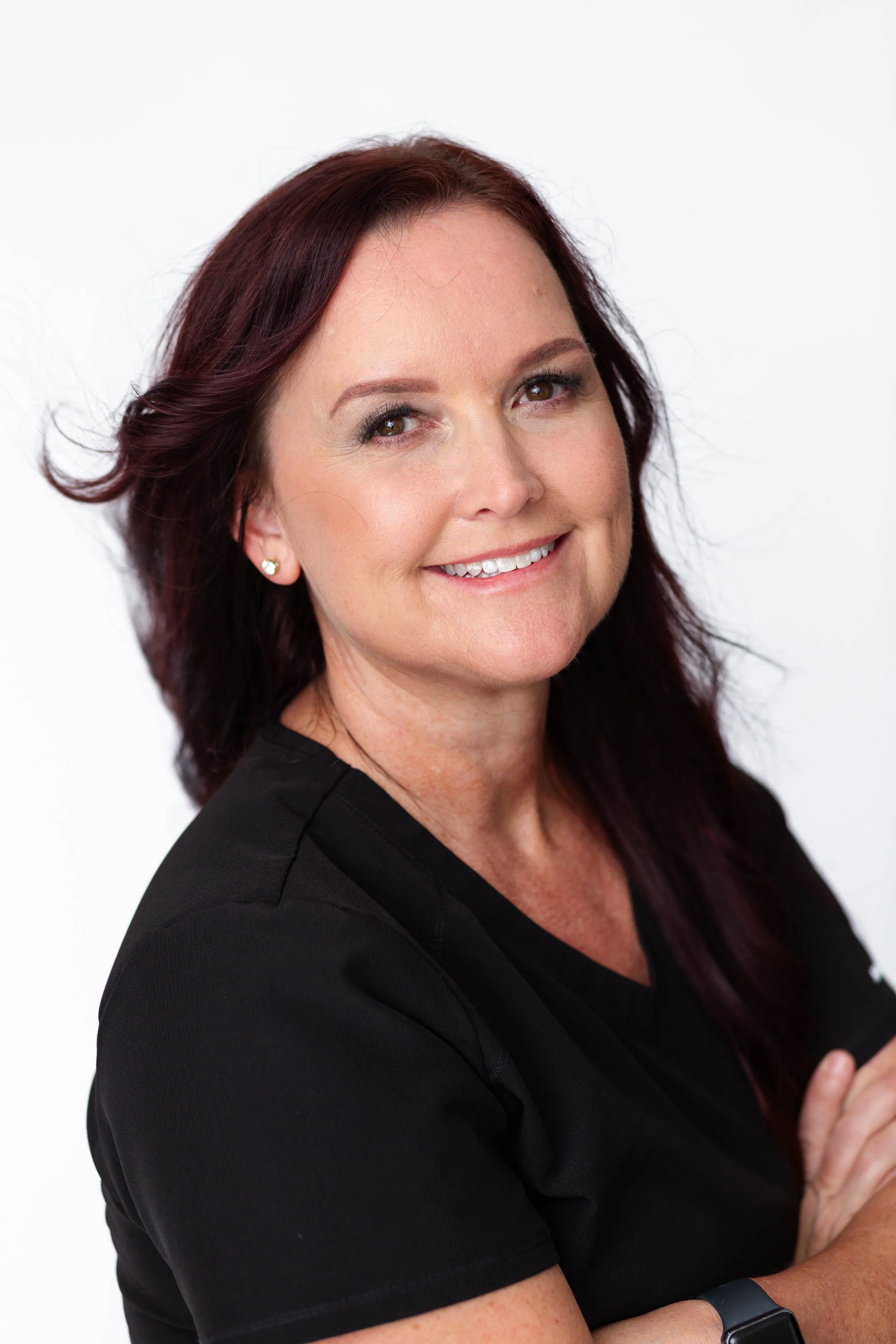 A smiling woman with shoulder-length auburn hair, wearing a black top and a smartwatch, crossing her arms against a plain white background.