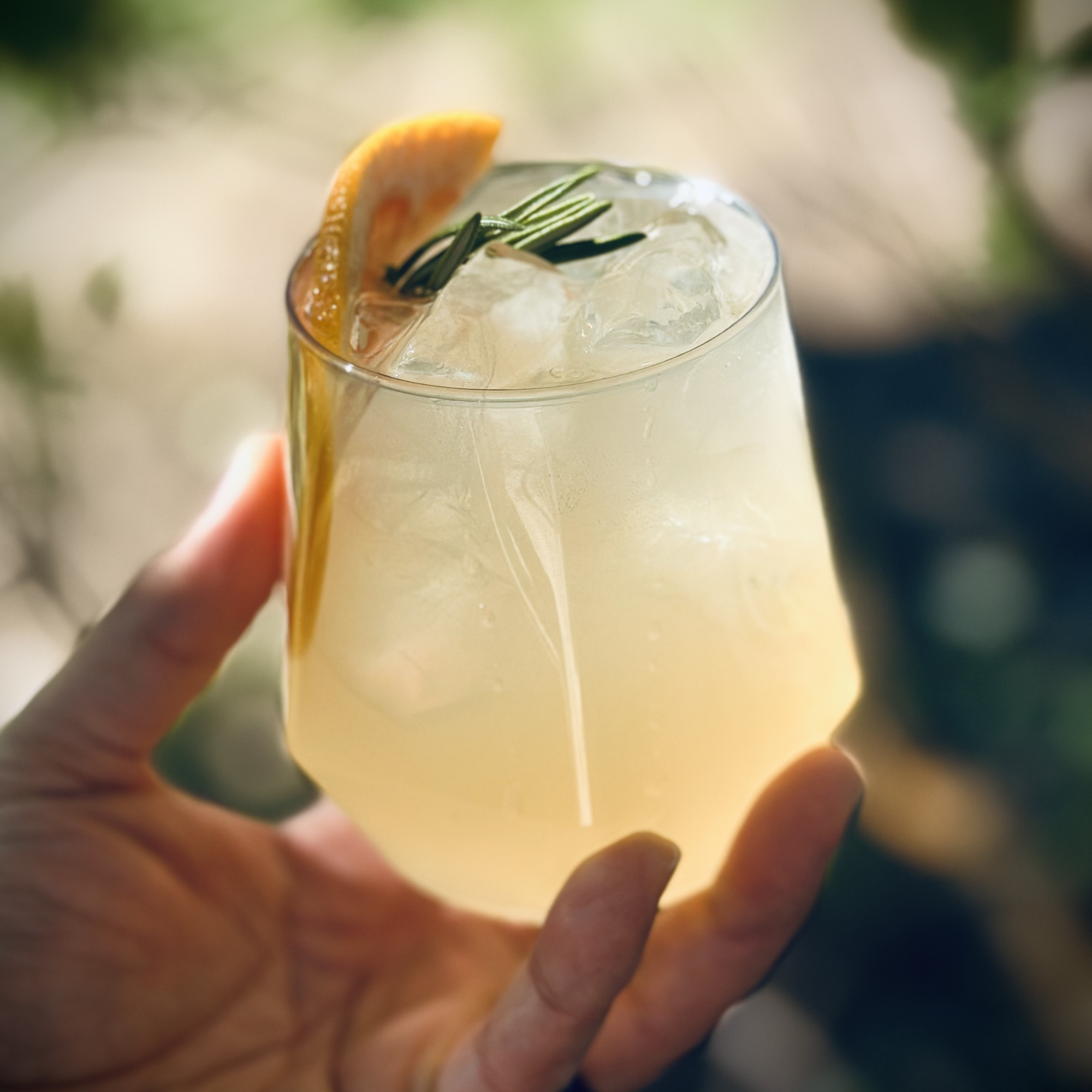 A handheld glass filled with a light-colored beverage, garnished with a lemon wedge and rosemary sprig, with ice cubes visible inside.