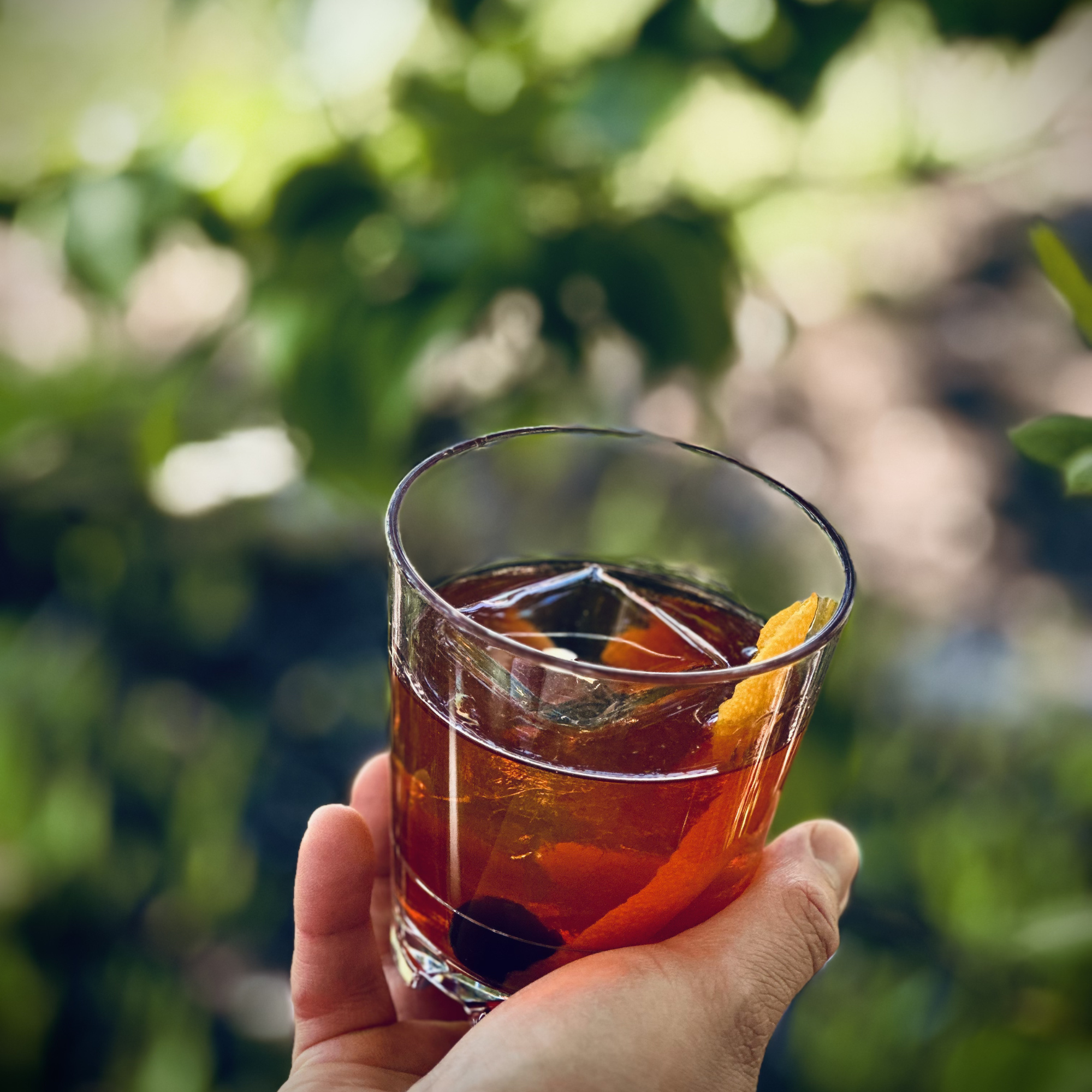 A close-up of a clear glass of dark iced tea with ice and an orange wedge, held by a person's hand against a blurred outdoor background with green foliage.