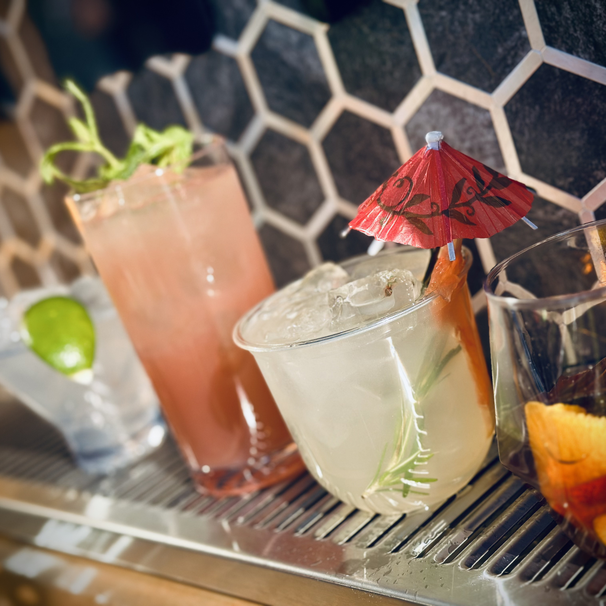 Close-up of a decorated paper cocktail umbrella in a glass, with a tiled dark background and a blurred plant in a pot.