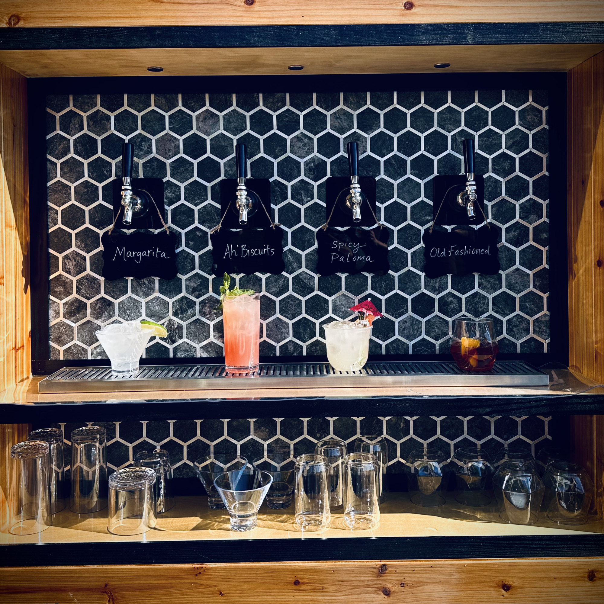 A bar setup with several empty glasses and cocktail drinks on a wooden shelf, with a black honeycomb-patterned tile background.