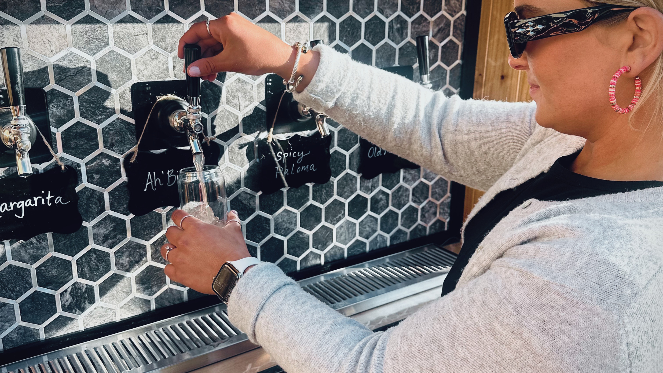 A woman filling a glass with beer from a tap at a bar or brewery, with a hexagonal tile wall in the background and chalkboard tags listing beer flavors.