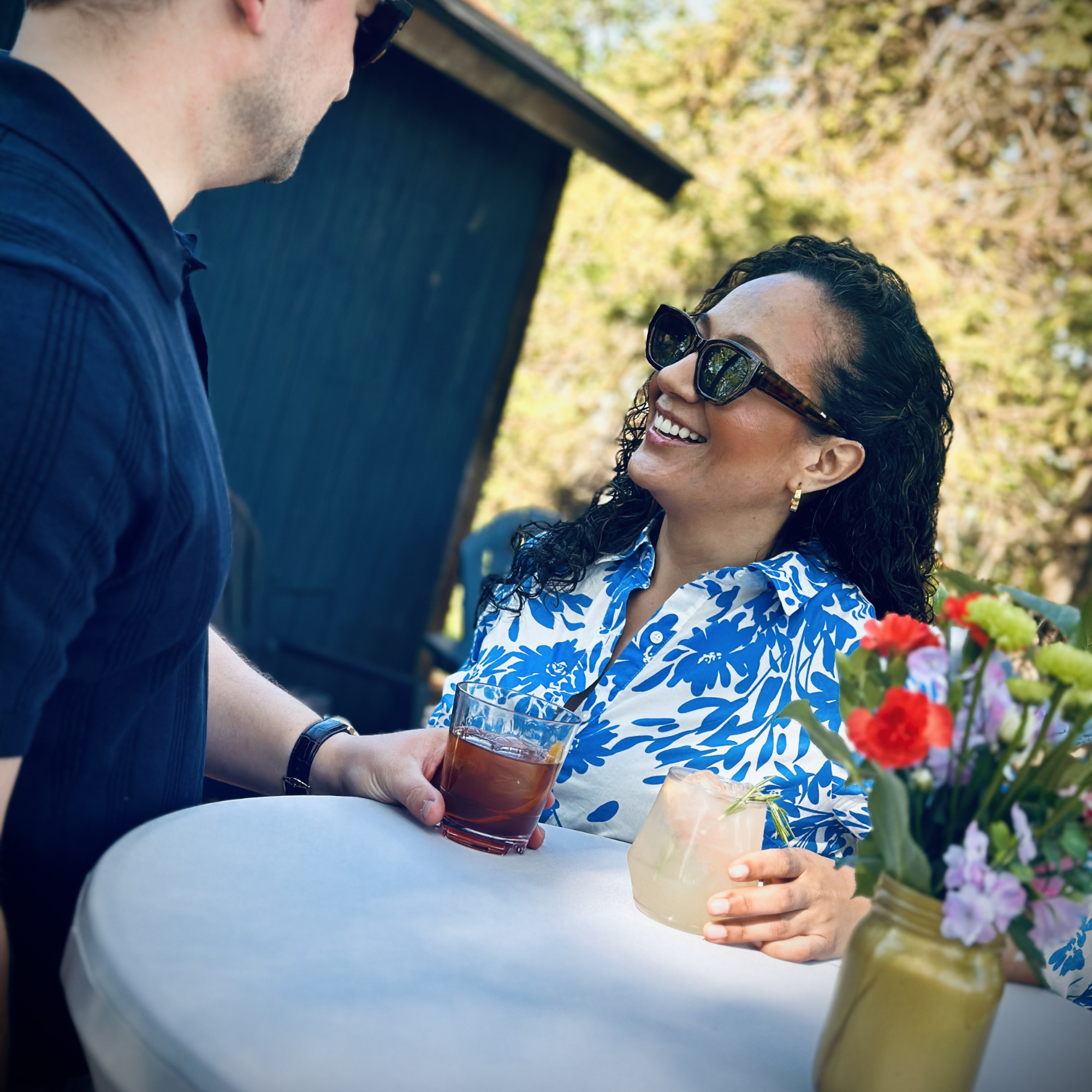 A man and a woman are outdoors on a sunny day. The man, wearing a dark shirt, is holding a drink, and the woman, wearing sunglasses and a blue and white patterned dress, is smiling. There are vibrant flowers in a jar on the table.