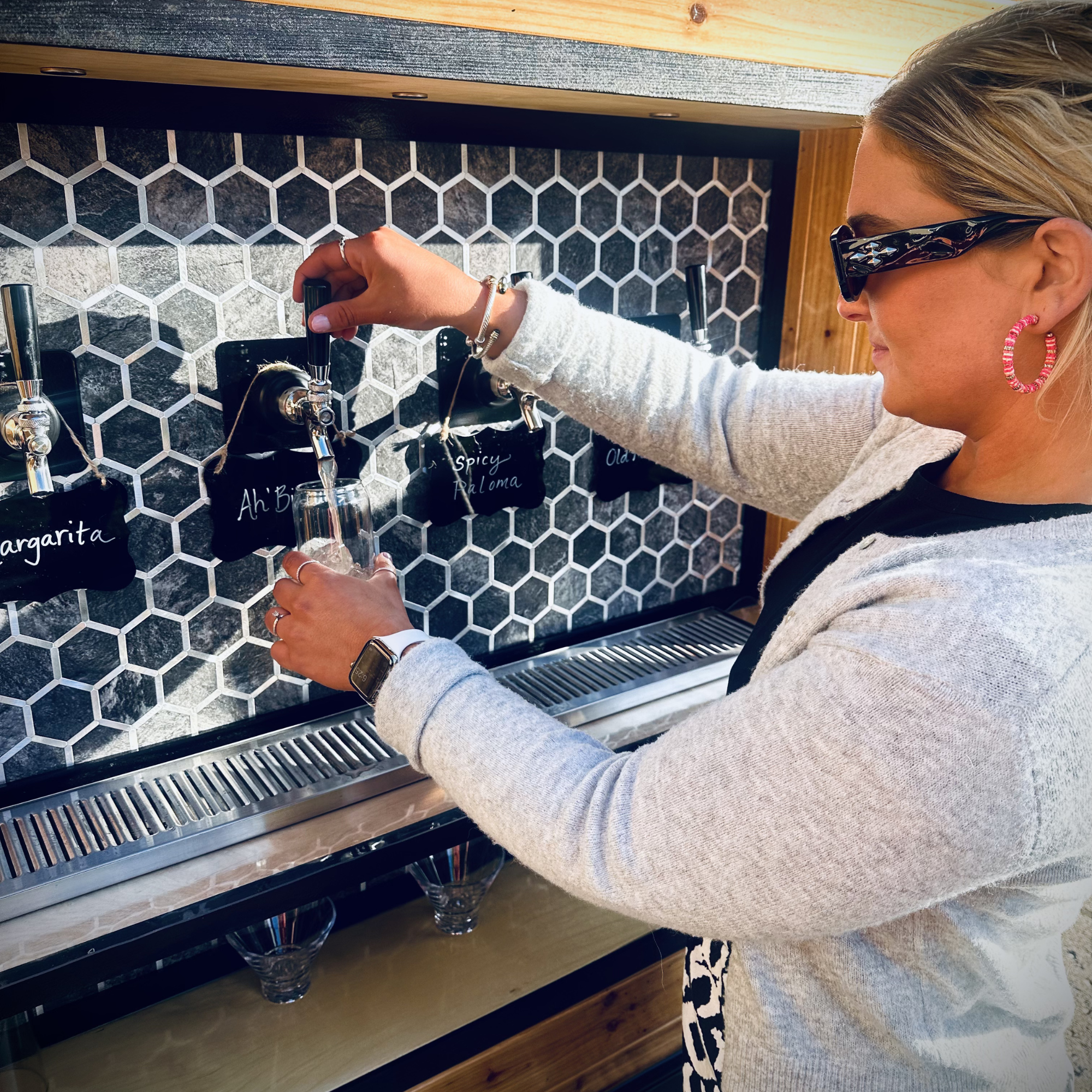 A woman with blonde hair wearing glasses and pink hoop earrings at a bar, pouring a drink from a tap. The bar has a honeycomb tile backsplash and a black chalkboard with drink names.