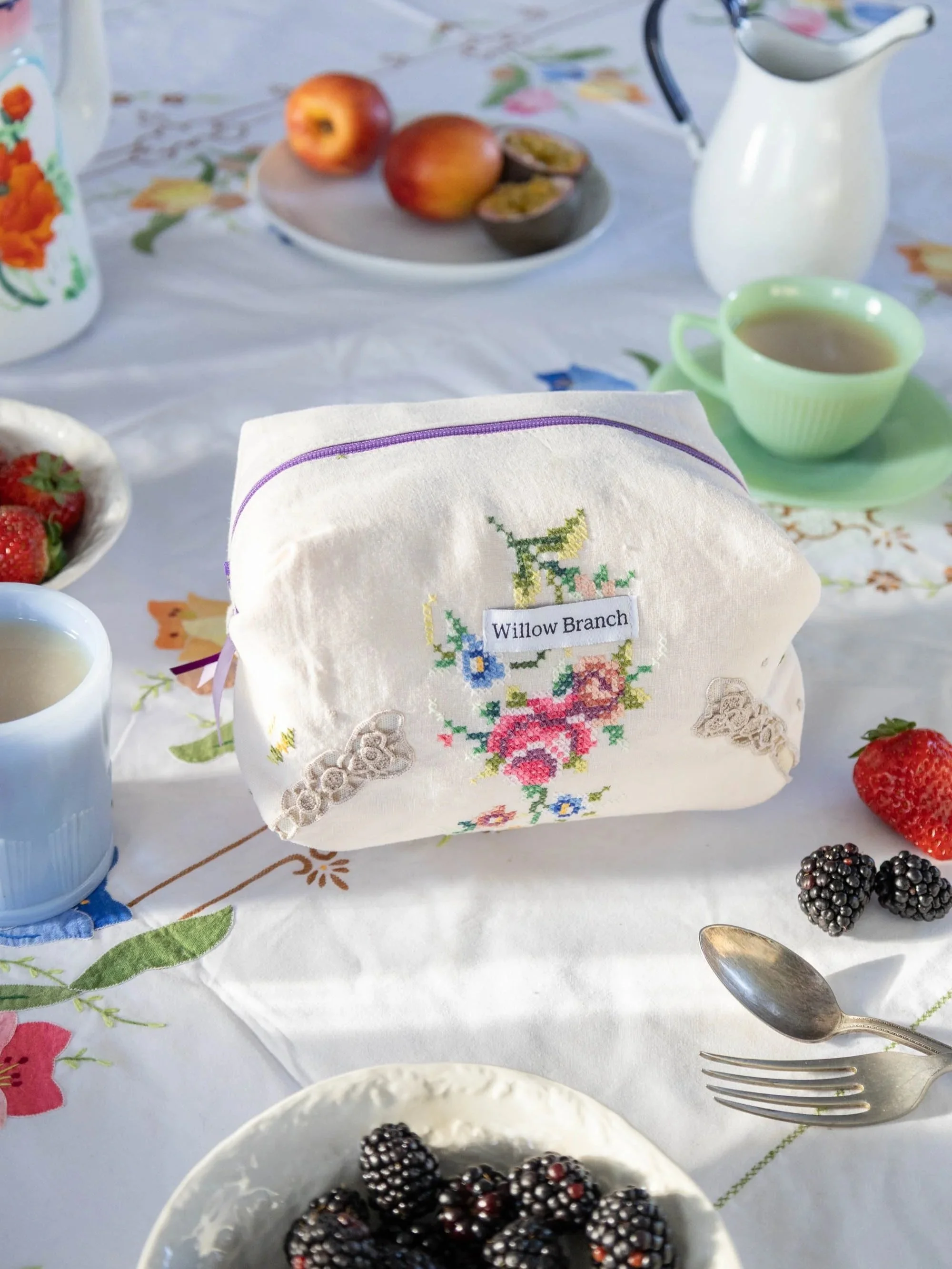 Willow Branch Boxy Makeup bag using fabric from a vintage embroidered table cloth. The makeup bag is sitting on a vintage table cloth with different fruits and cups of tea around.