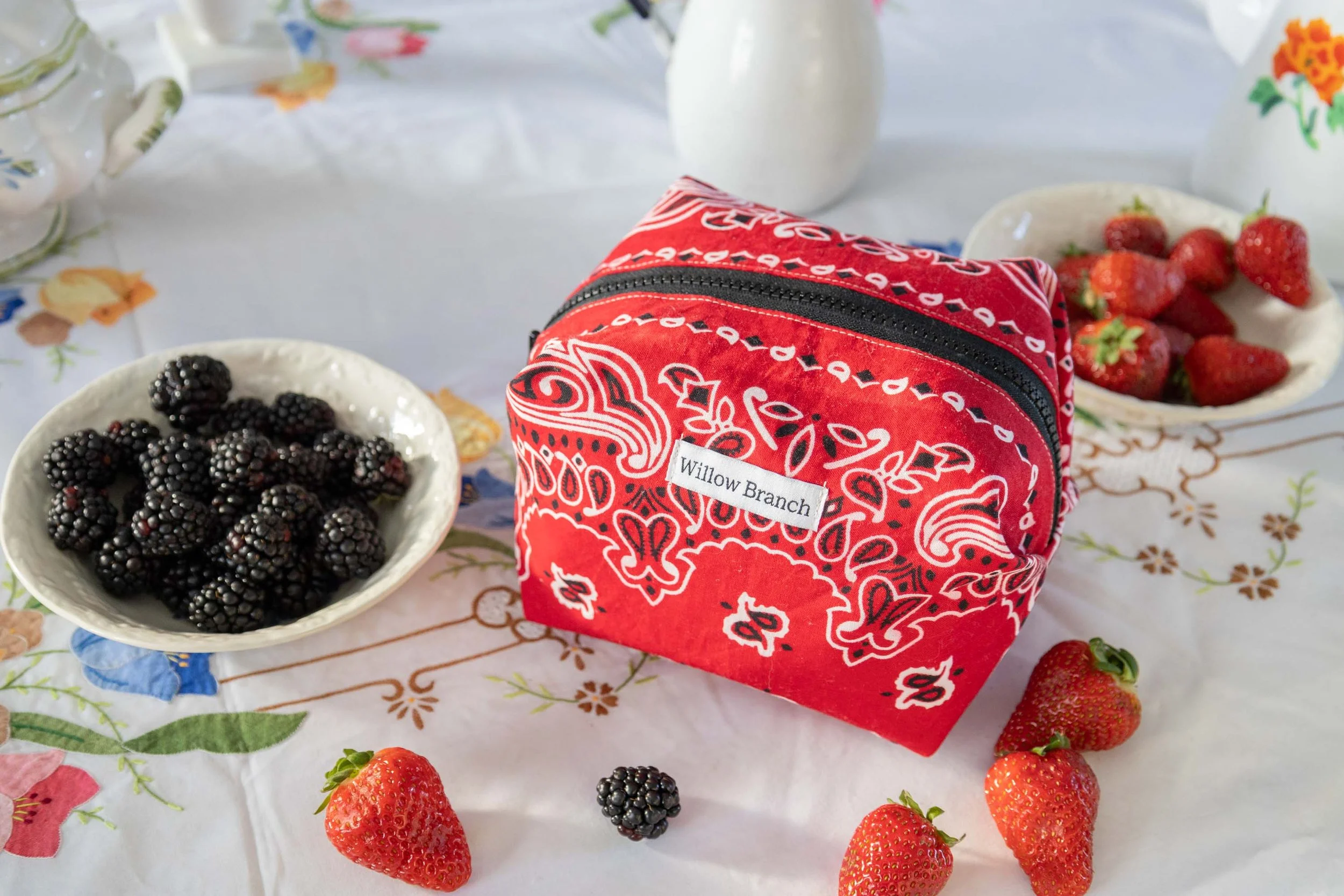 Red and white bandana print boxy makeup bag sitting on a vintage table cloth with a bowl of black berries on the side and strawberries and blackberries scattered on the table