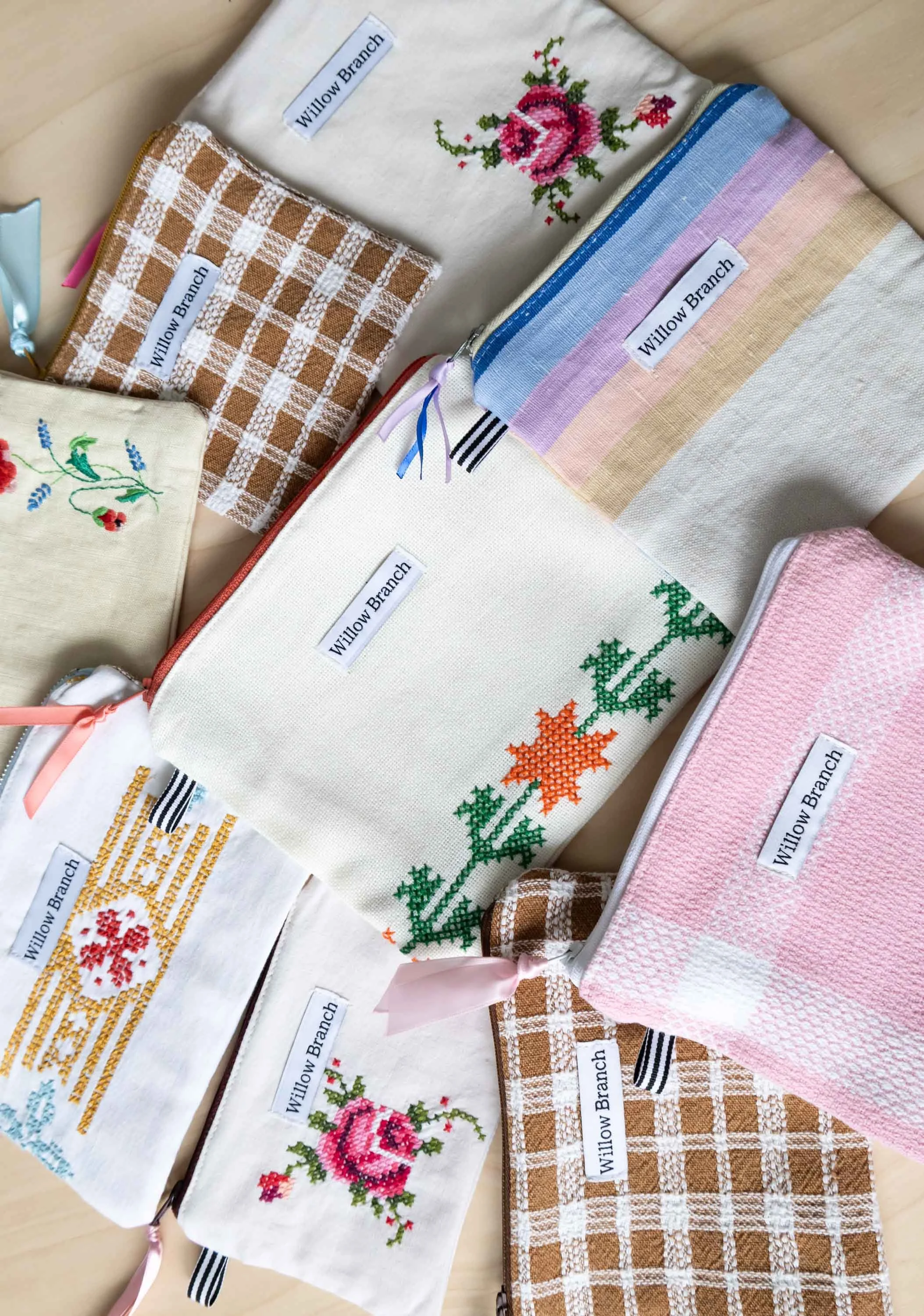 A flat-lay of multiple flat pouches with different colors, textures, and embroidery designs. Each one displaying the signature Willow Branch label, and black and white ribbon tab.