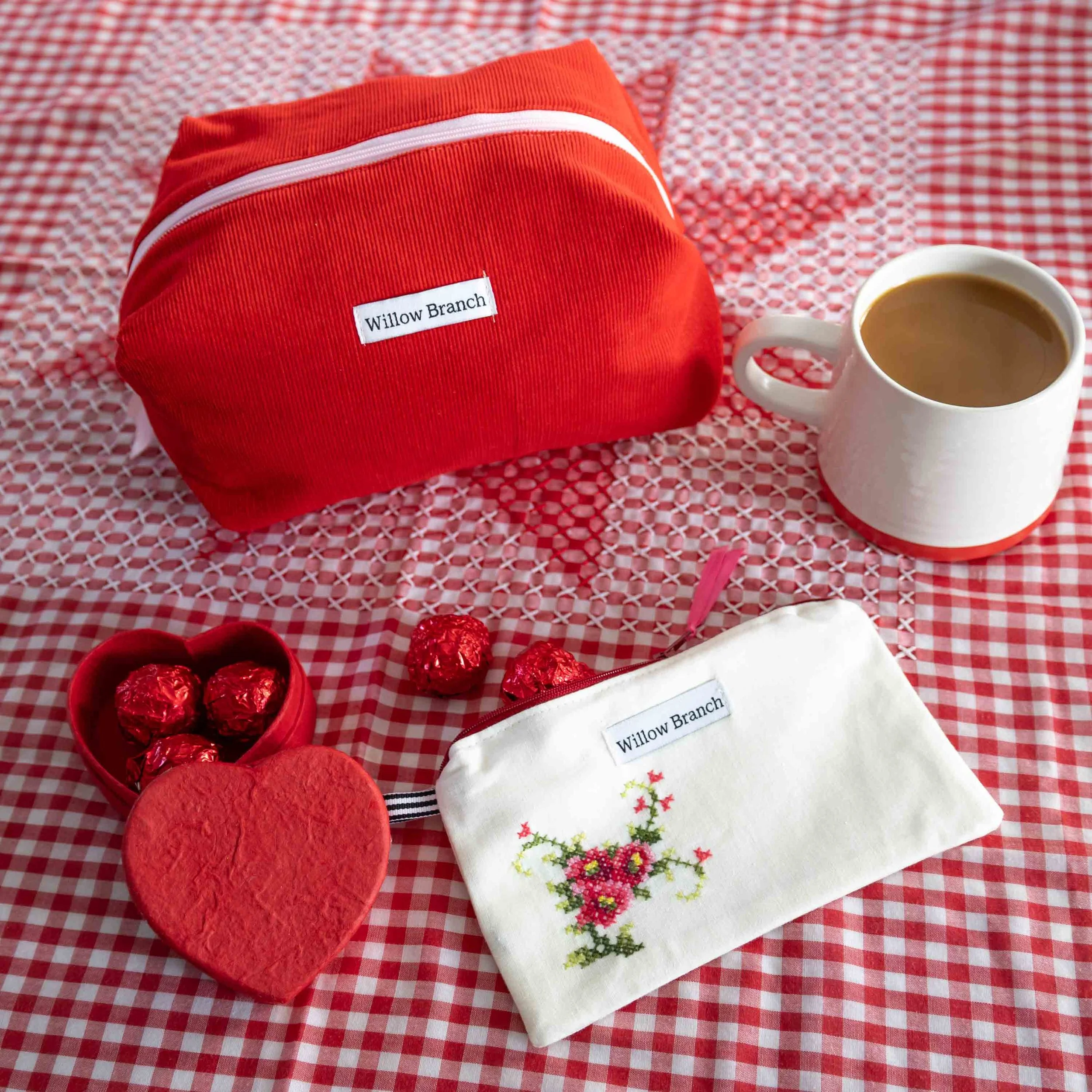 Vintage Embroidery Flat Pouch with a Rose Motif and Red Corduroy Makeup Bag on a Red Gingham table cloth. In the frame there is a red heart box with red foil covered chocolates and a cup of coffee.