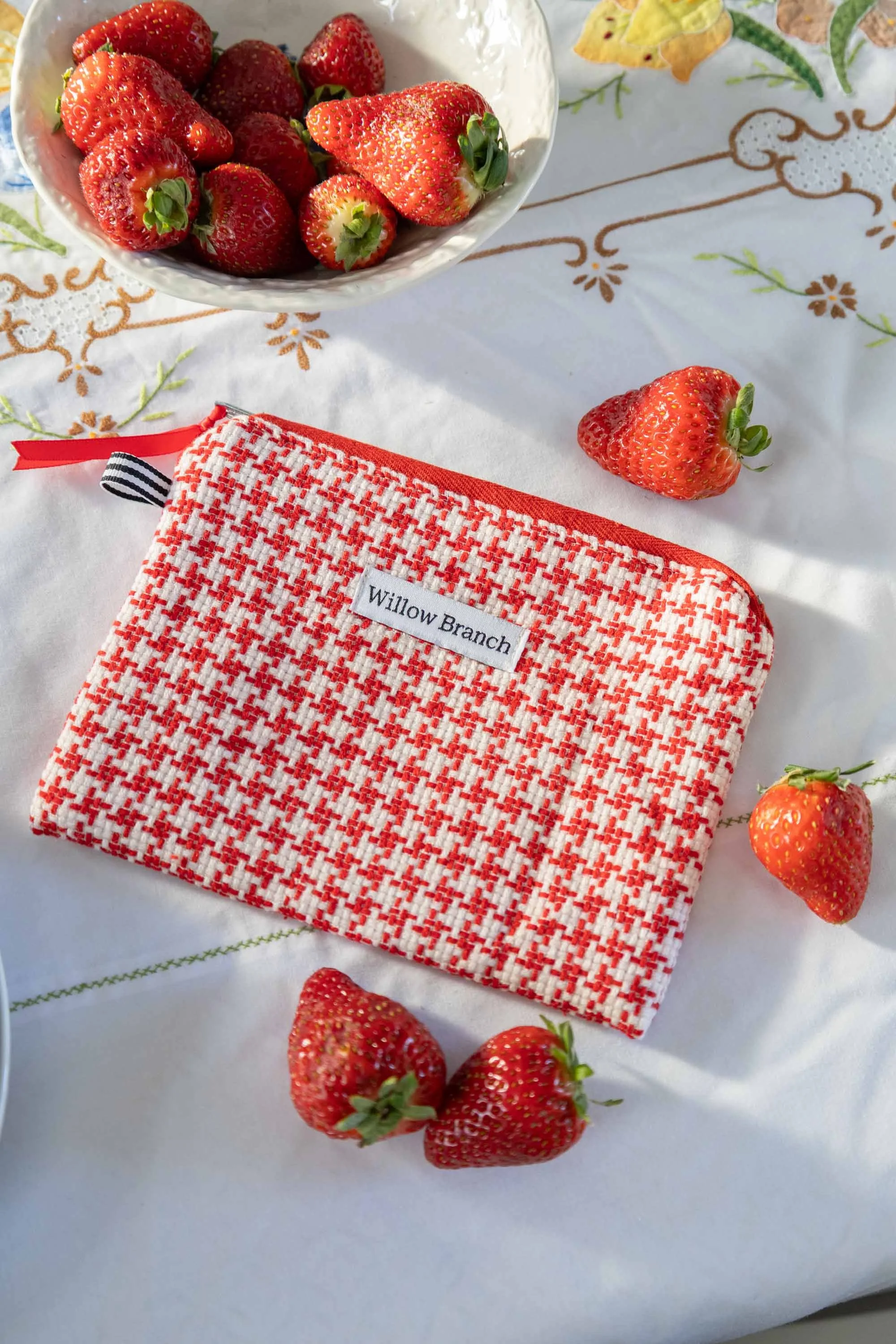 Red and white flat pouch with beautiful morning light hitting it on a table with strawberries strewn about and a bowl of strawberries on the side all sitting n top of a vintage table cloth