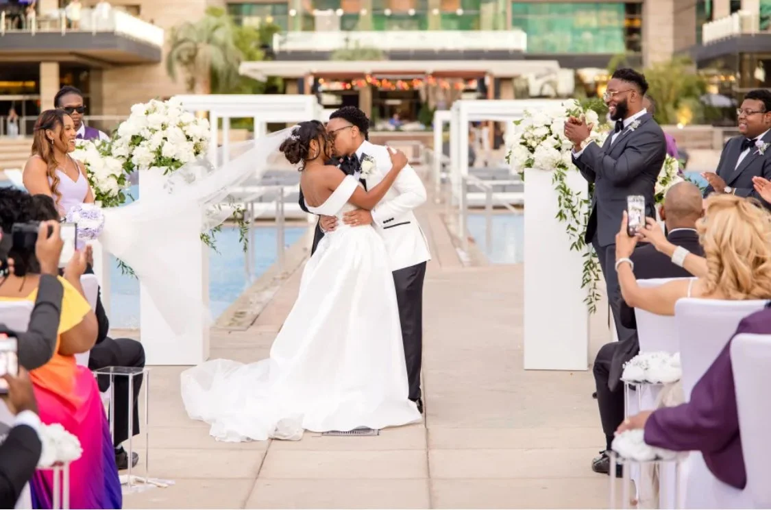  couple kissing at their wedding ceremony outdoors, surrounded by friends and family, with floral decorations and city buildings in the background. 