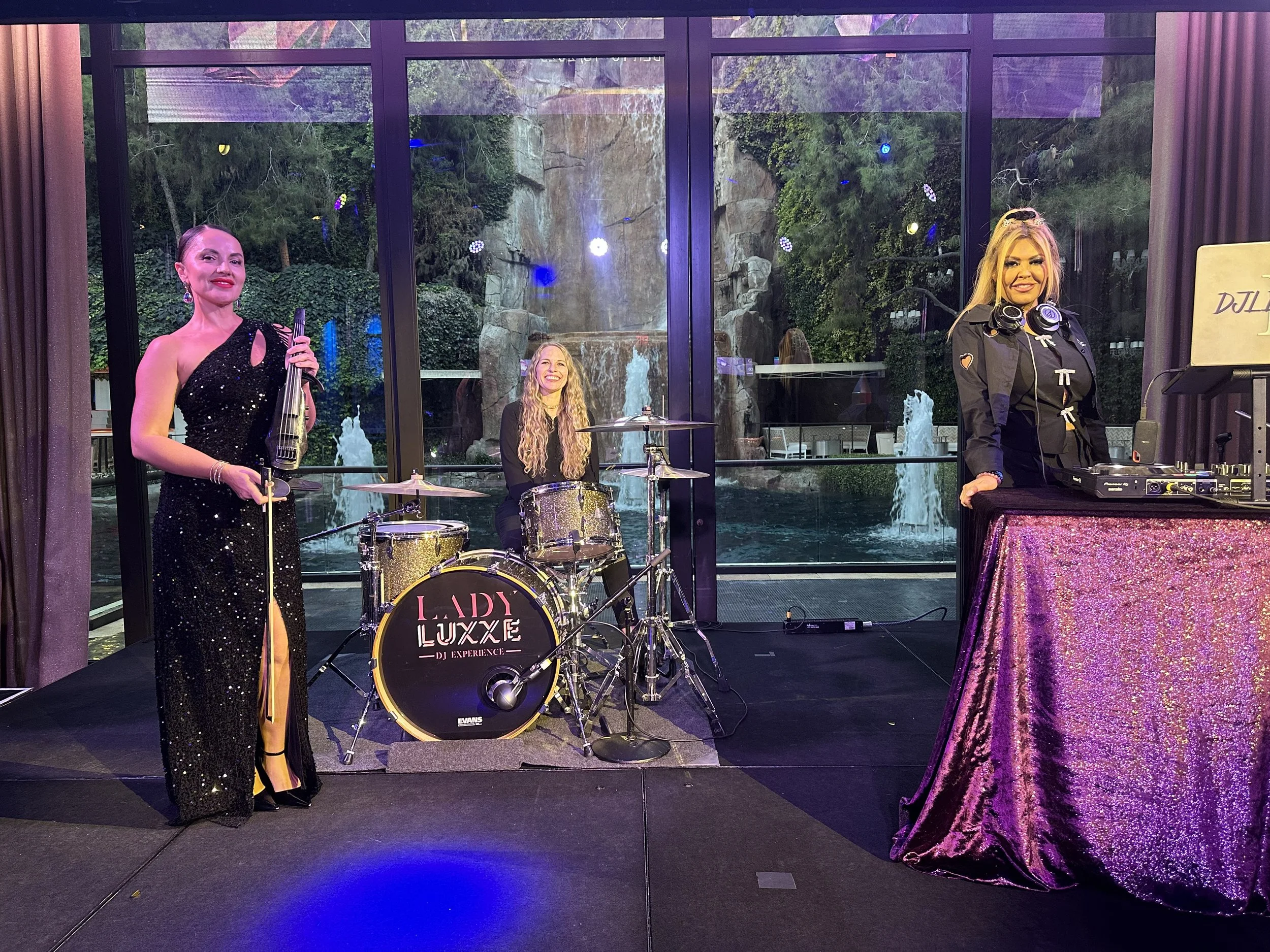 Three women performing music indoors with a large window showing a waterfall and trees outside. One woman stands holding a bass guitar, another sits behind a drum kit labeled Lady Luxxe DJ Experience, and the third woman stands behind a DJ setup with