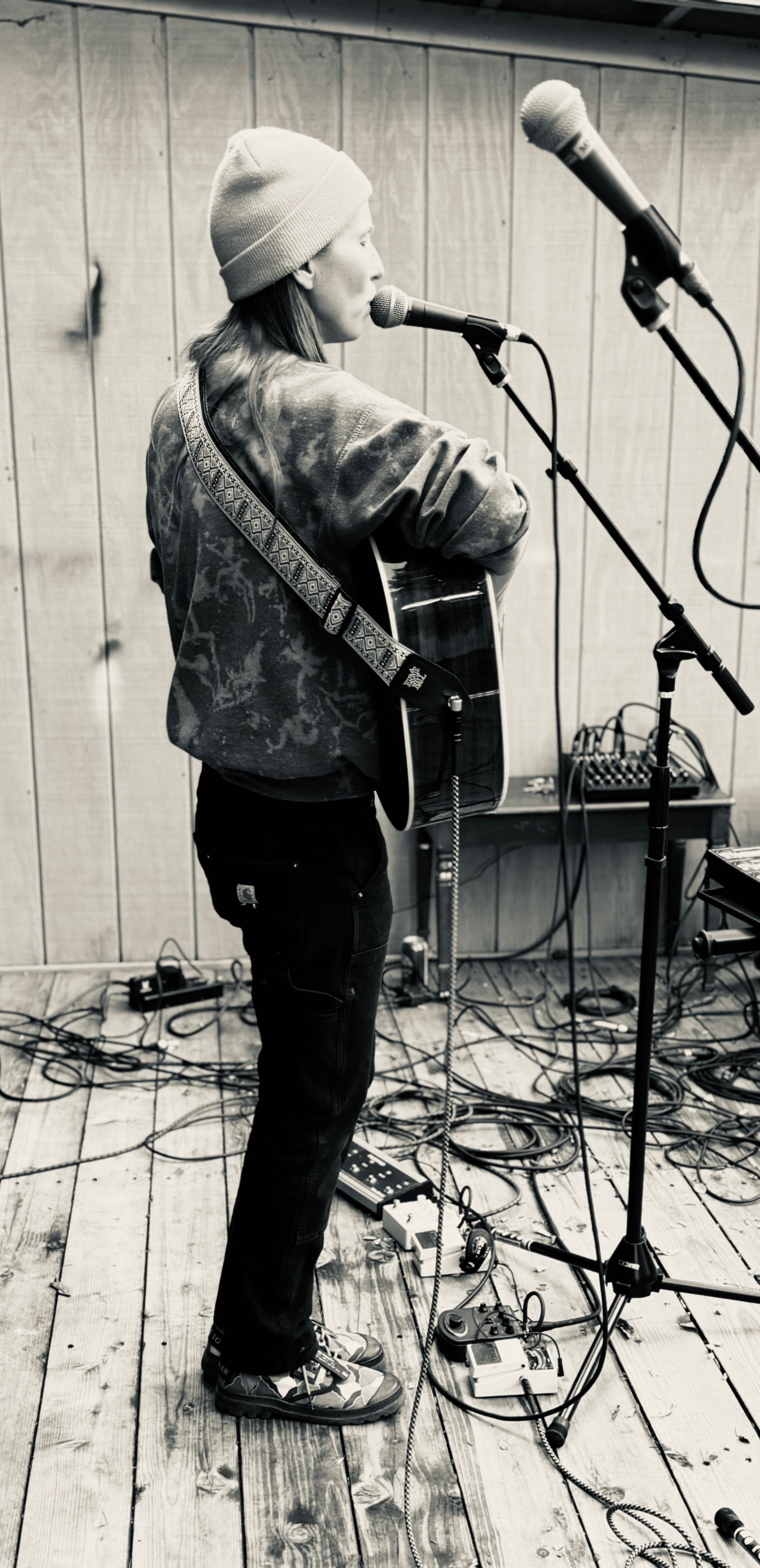 A woman wearing a beanie hat and a patterned jacket is playing an acoustic guitar into a microphone in a music studio with wooden walls. There are multiple microphones and audio equipment on the floor and a music stand in the background.