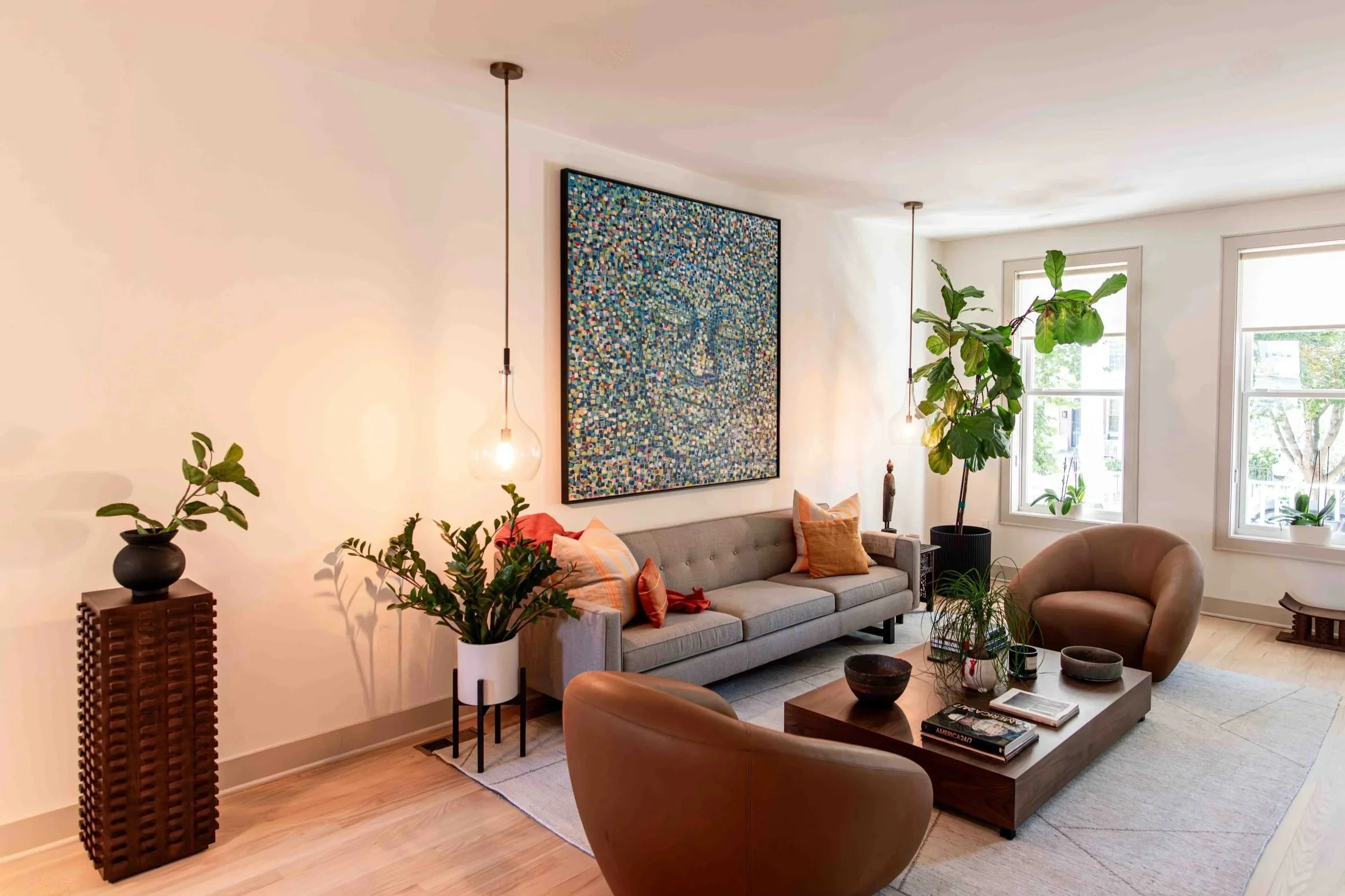 Bright living room with a light gray sofa, a round tan armchair, and a rectangular wooden coffee table. Potted plants are near the windows and on the side tables. Two hanging lights with exposed bulbs are above the sofa. There is a large framed abstr