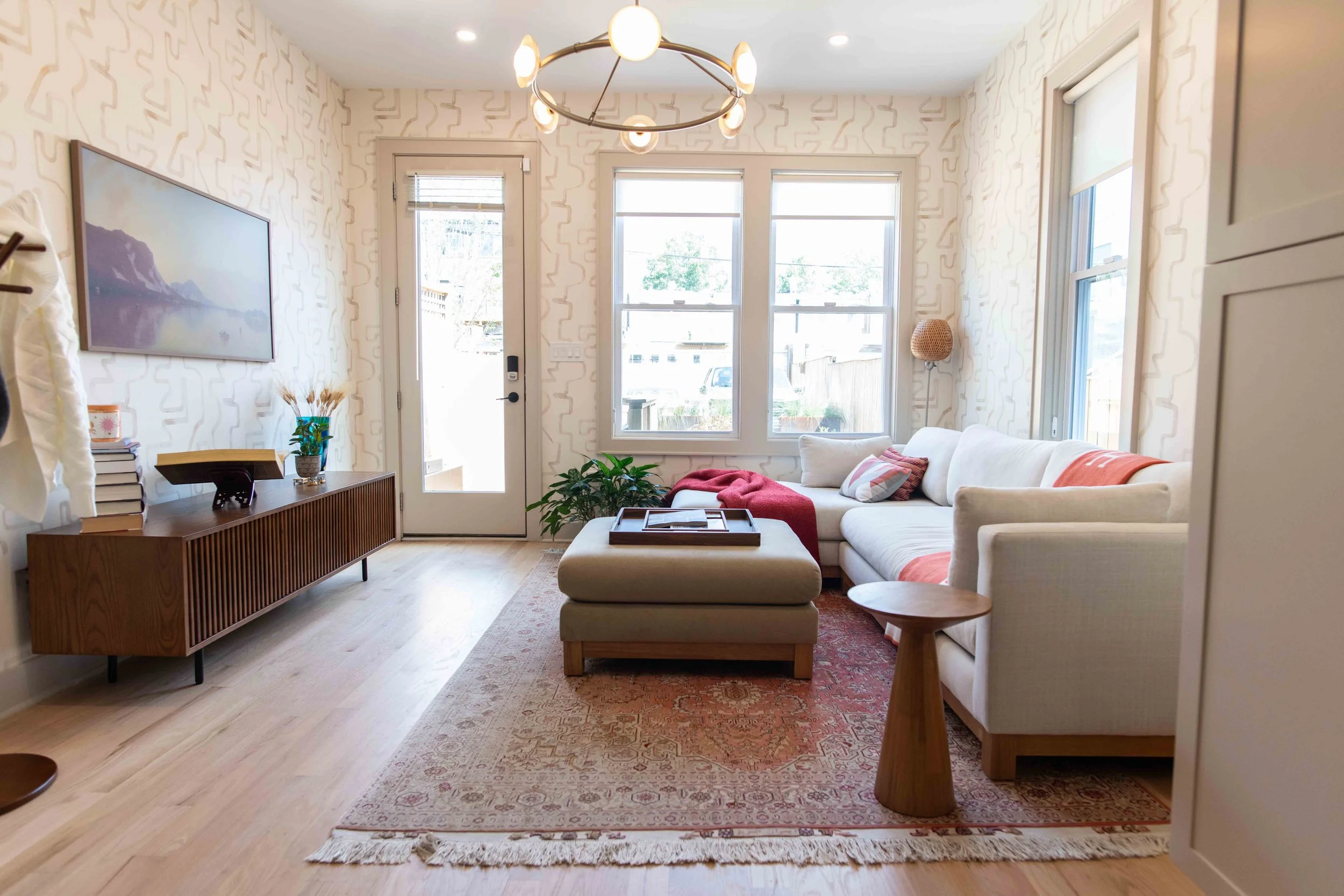 Bright living room with cream sectional sofa, wooden side table, patterned rug, wall-mounted television, and large windows.