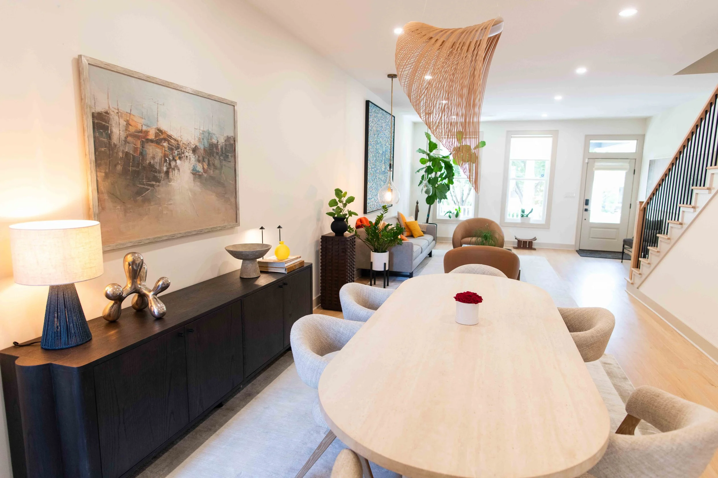 Interior view of a modern, bright living and dining area with a light-colored wood table, beige chairs, a gray sofa with yellow and orange pillows, large windows, and a staircase with black balusters. Decor includes a large abstract painting, a black